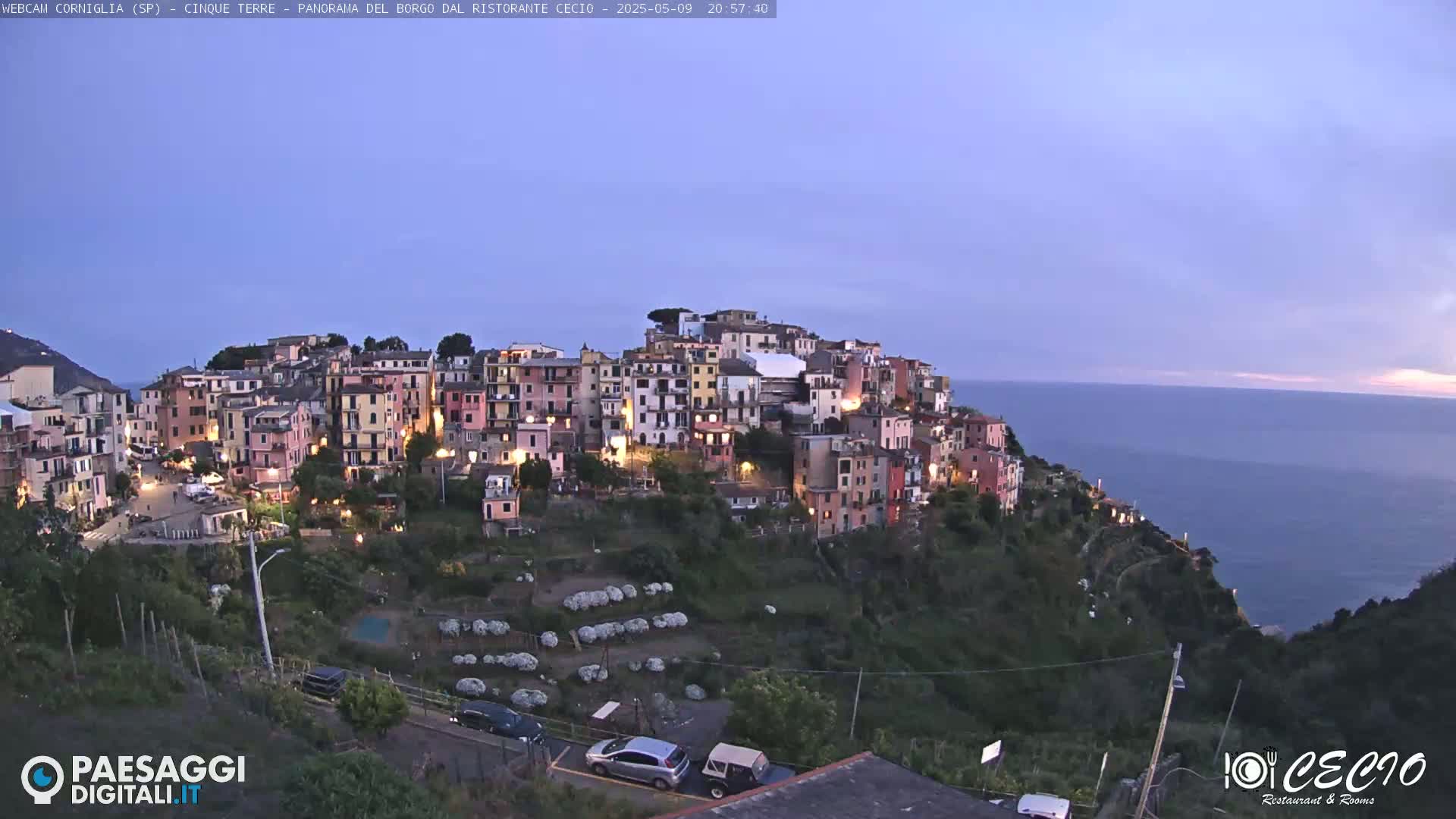 A colorful hillside village at twilight overlooks a calm ocean under a clear, dusky sky.