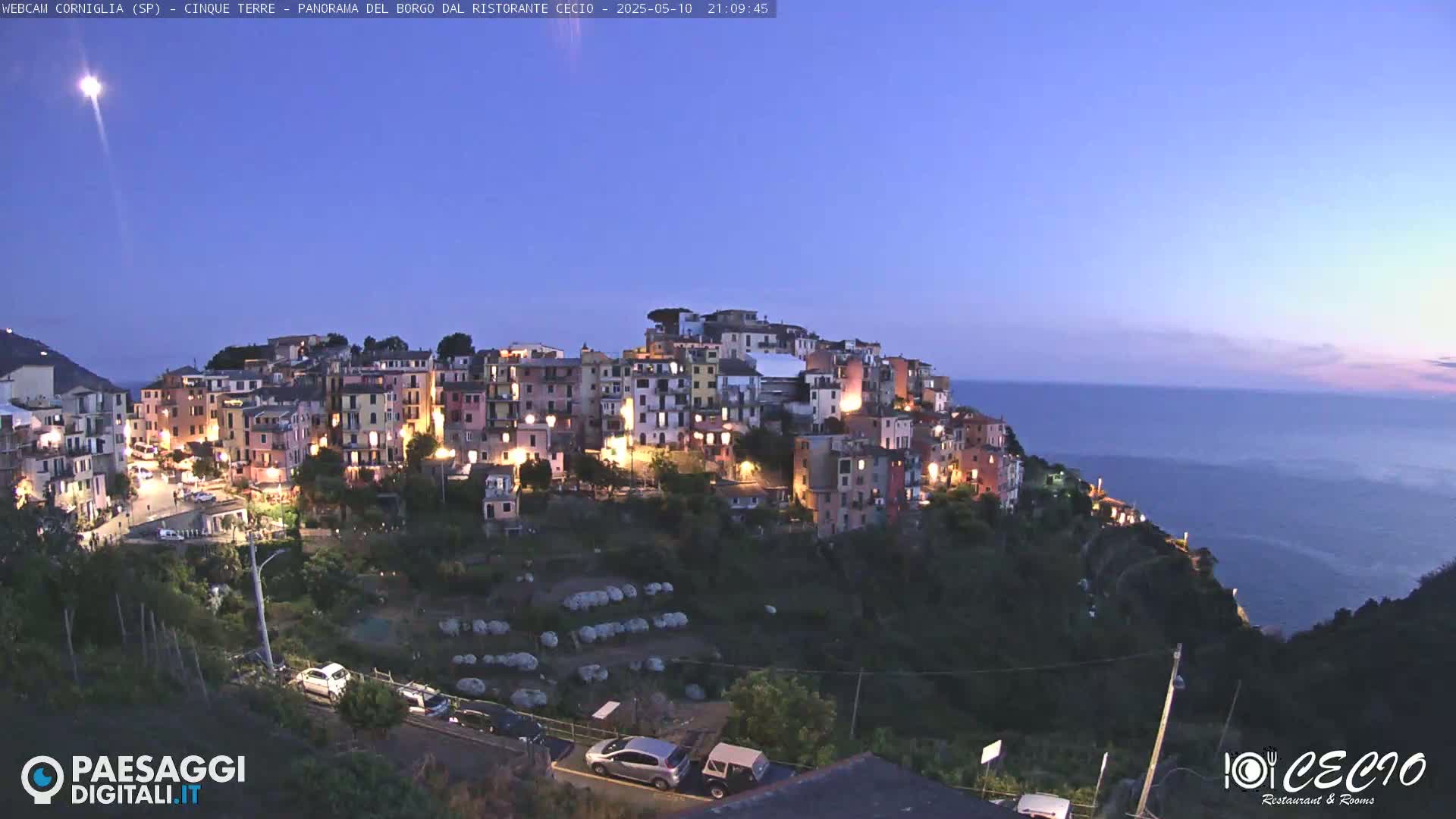 A brightly lit coastal village sits on a cliff overlooking a calm, dark ocean under a twilight sky.