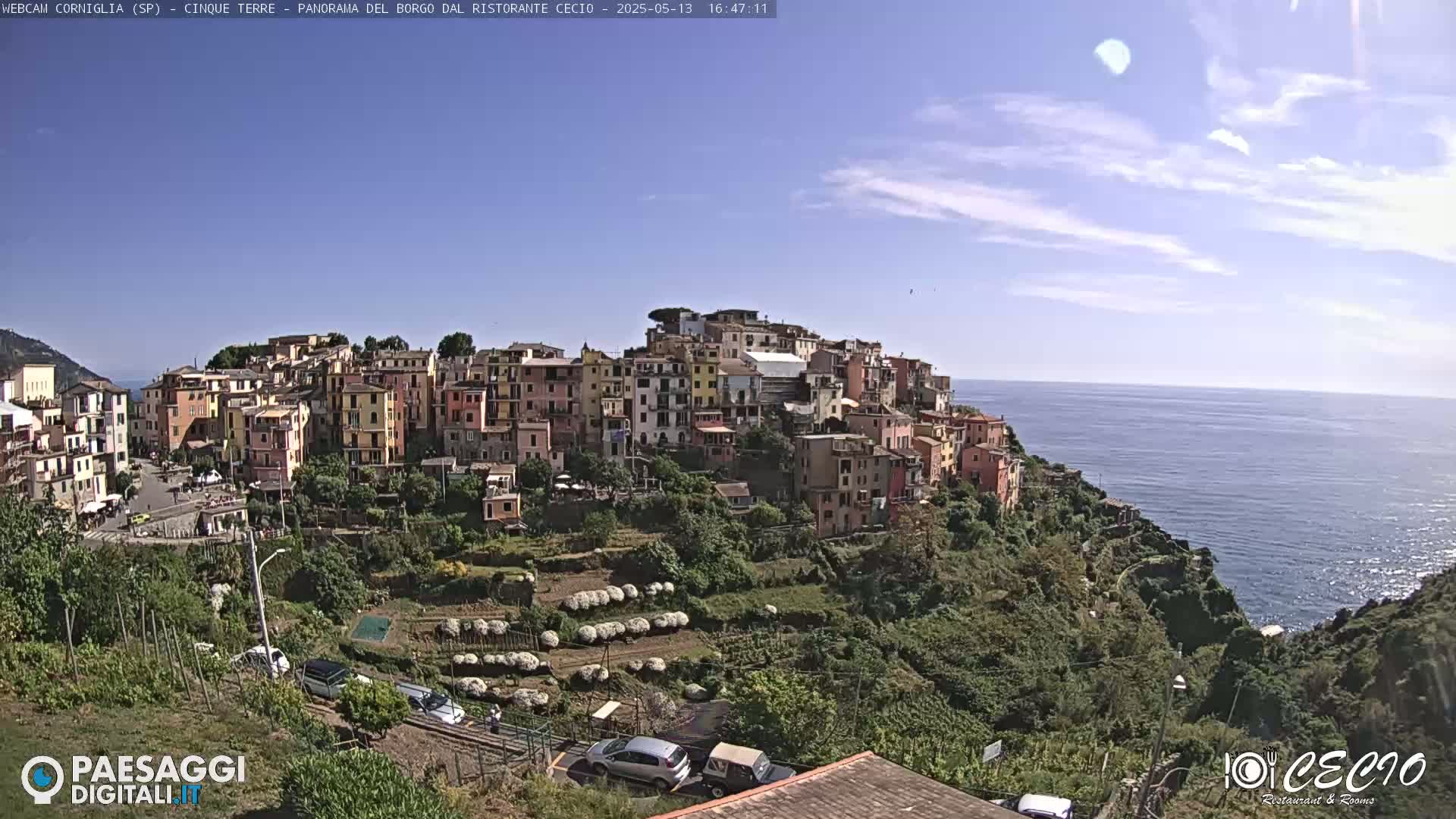 A colorful village clings to a hillside overlooking a calm ocean under a mostly clear, sunny sky.