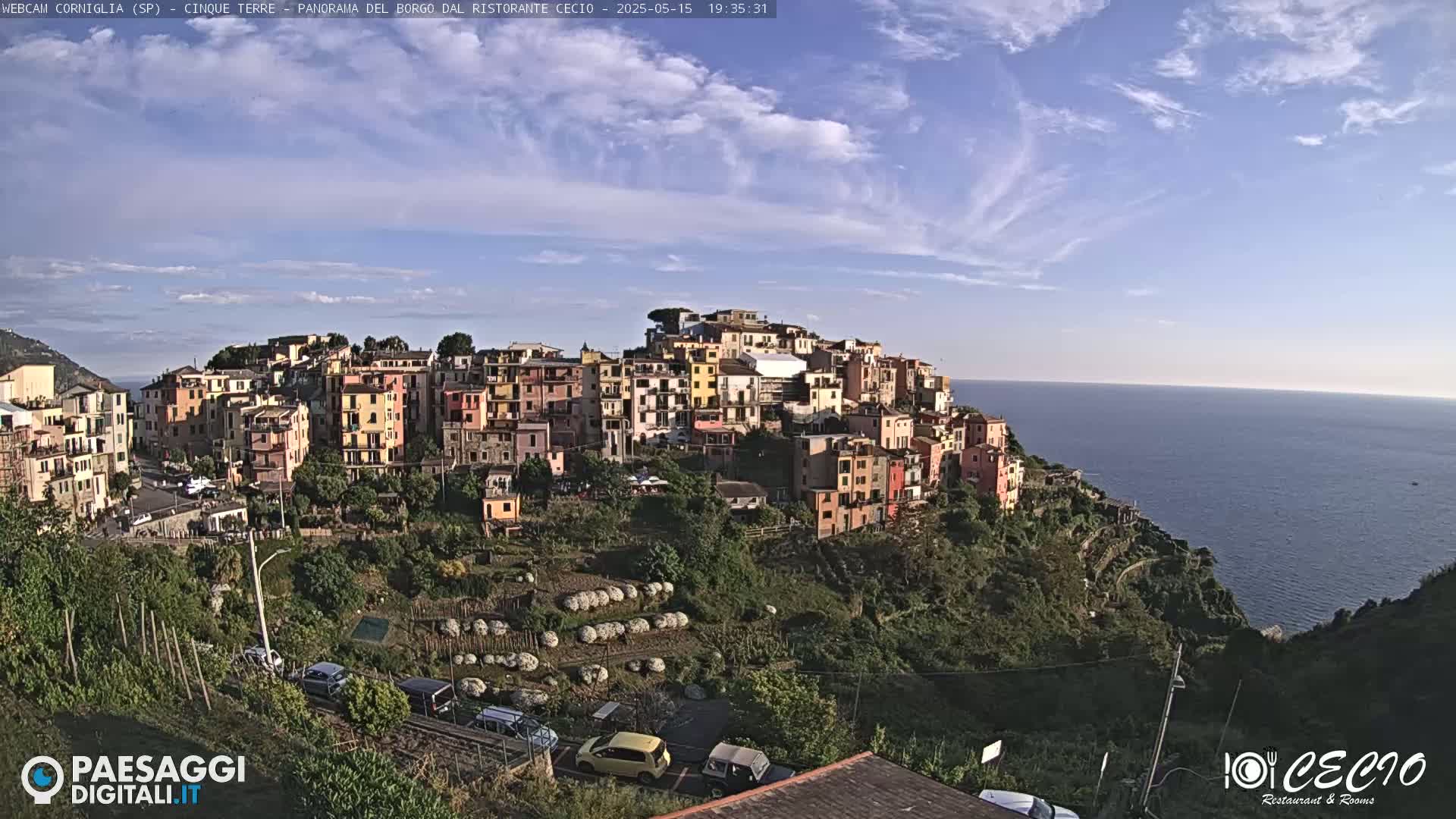 A picturesque coastal town with colorful buildings clings to a hillside overlooking a calm ocean under a mostly clear, bright sky.