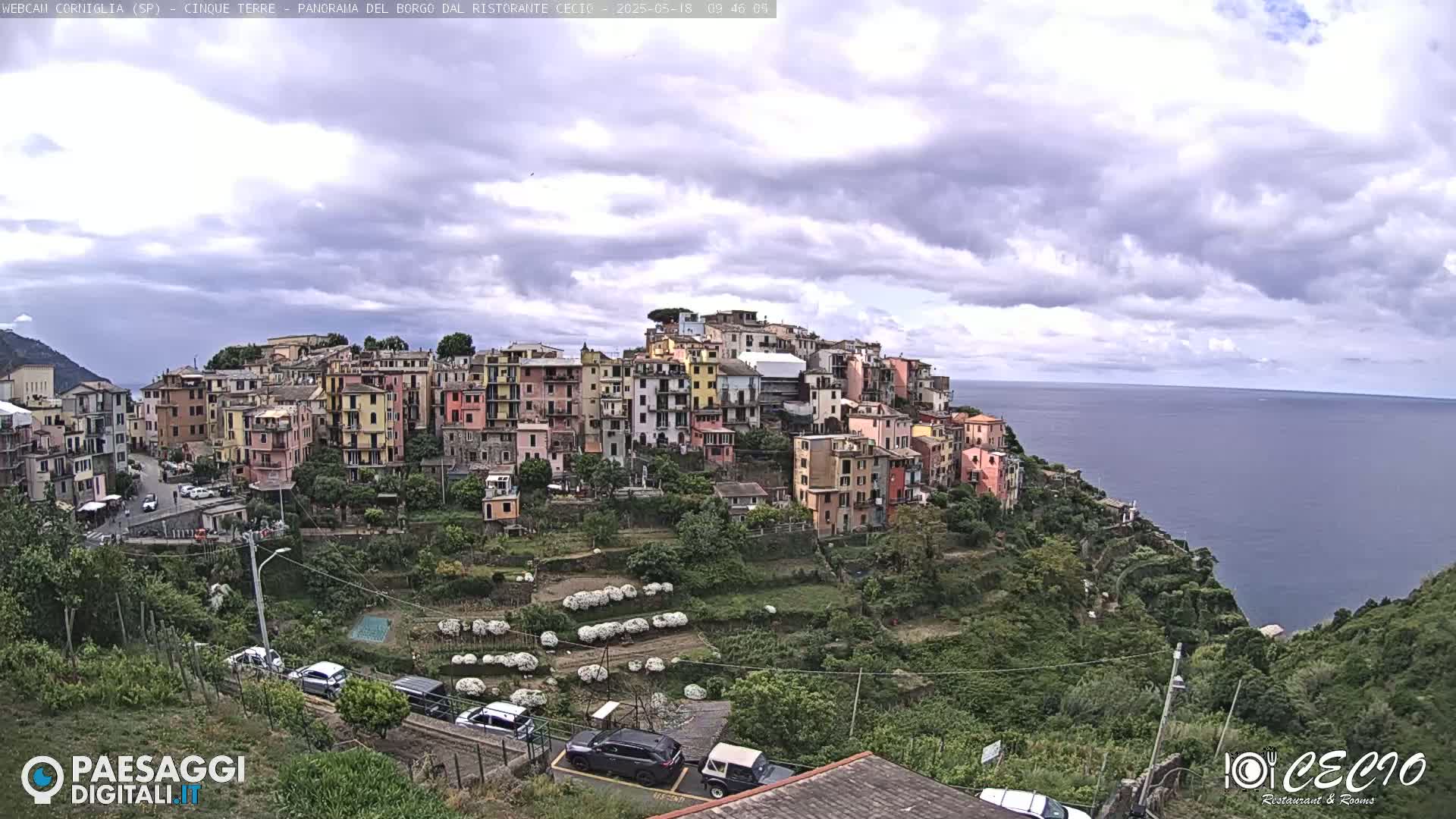 A colorful village clings to a hillside overlooking a calm sea under a partly cloudy sky.