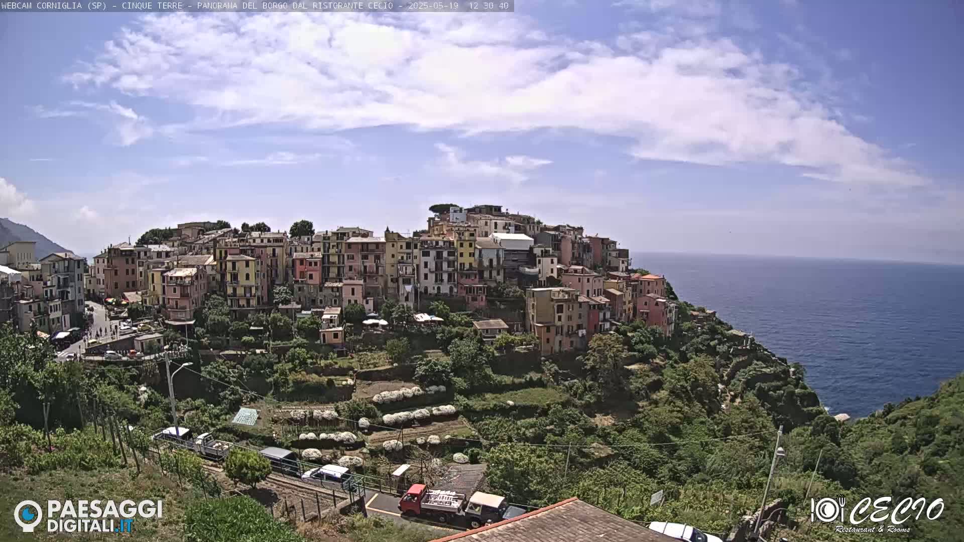 A picturesque coastal town, built on a hillside overlooking the sea under a partly cloudy sky, features colorful buildings and terraced gardens.