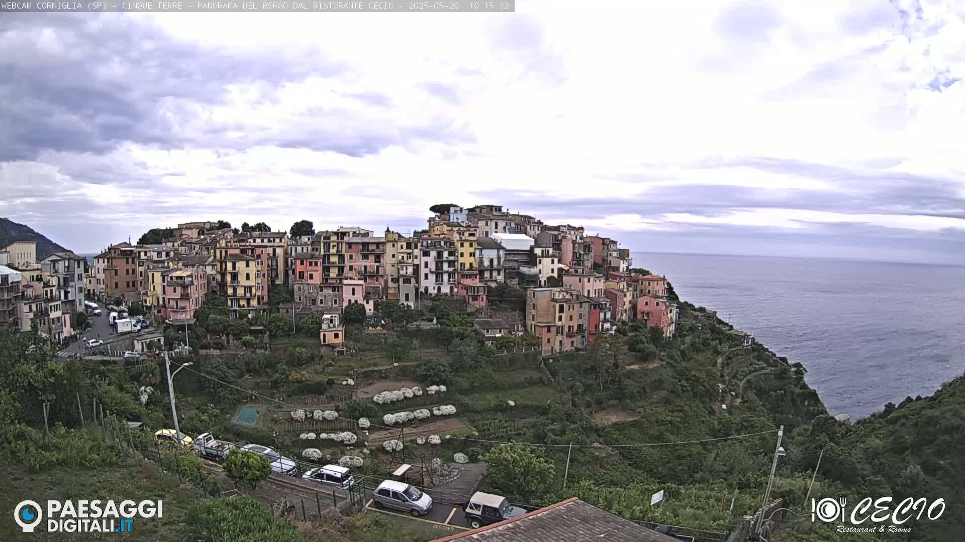 A coastal village of colorful buildings clings to a hillside overlooking a calm sea under a mostly cloudy sky.