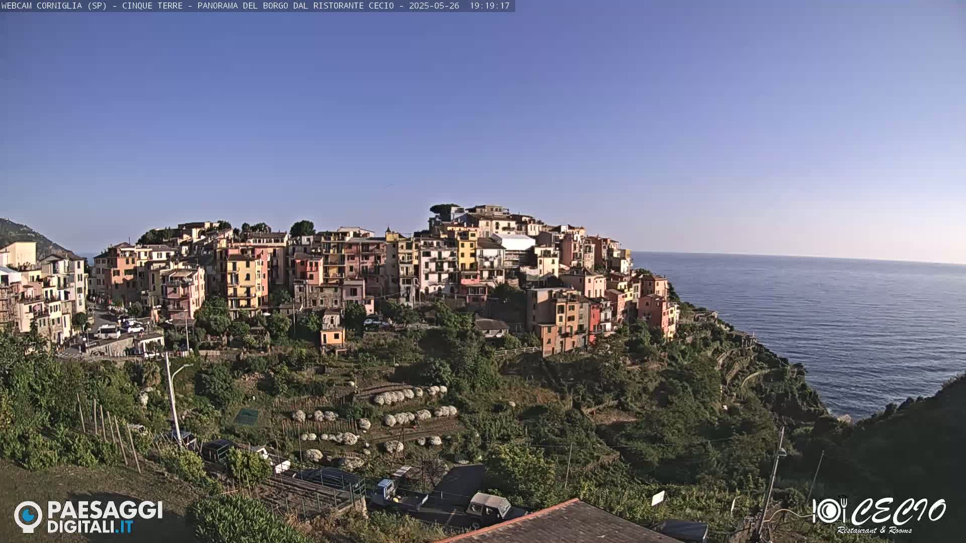 A colorful village clings to a hillside overlooking a calm blue sea under a clear blue sky.