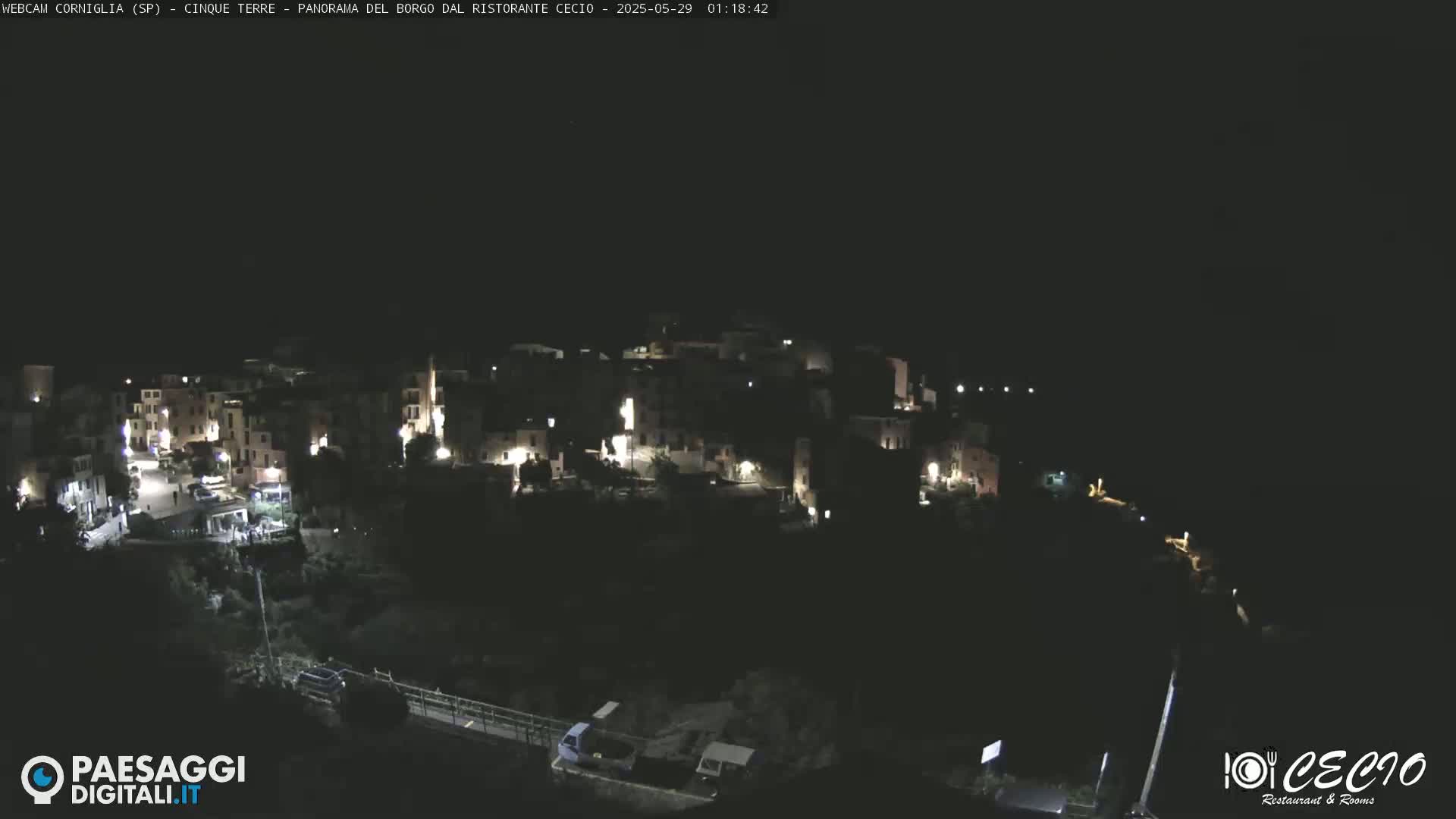 A nighttime, high-angle view shows a small, dimly lit village nestled on a dark hillside.