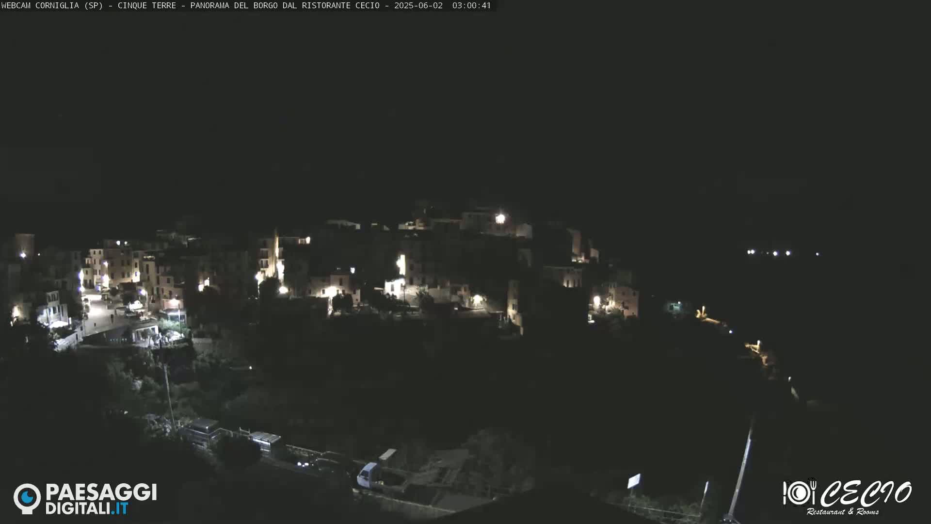 A nighttime, aerial view shows a small, hilly town with scattered lights, nestled in a dark landscape.