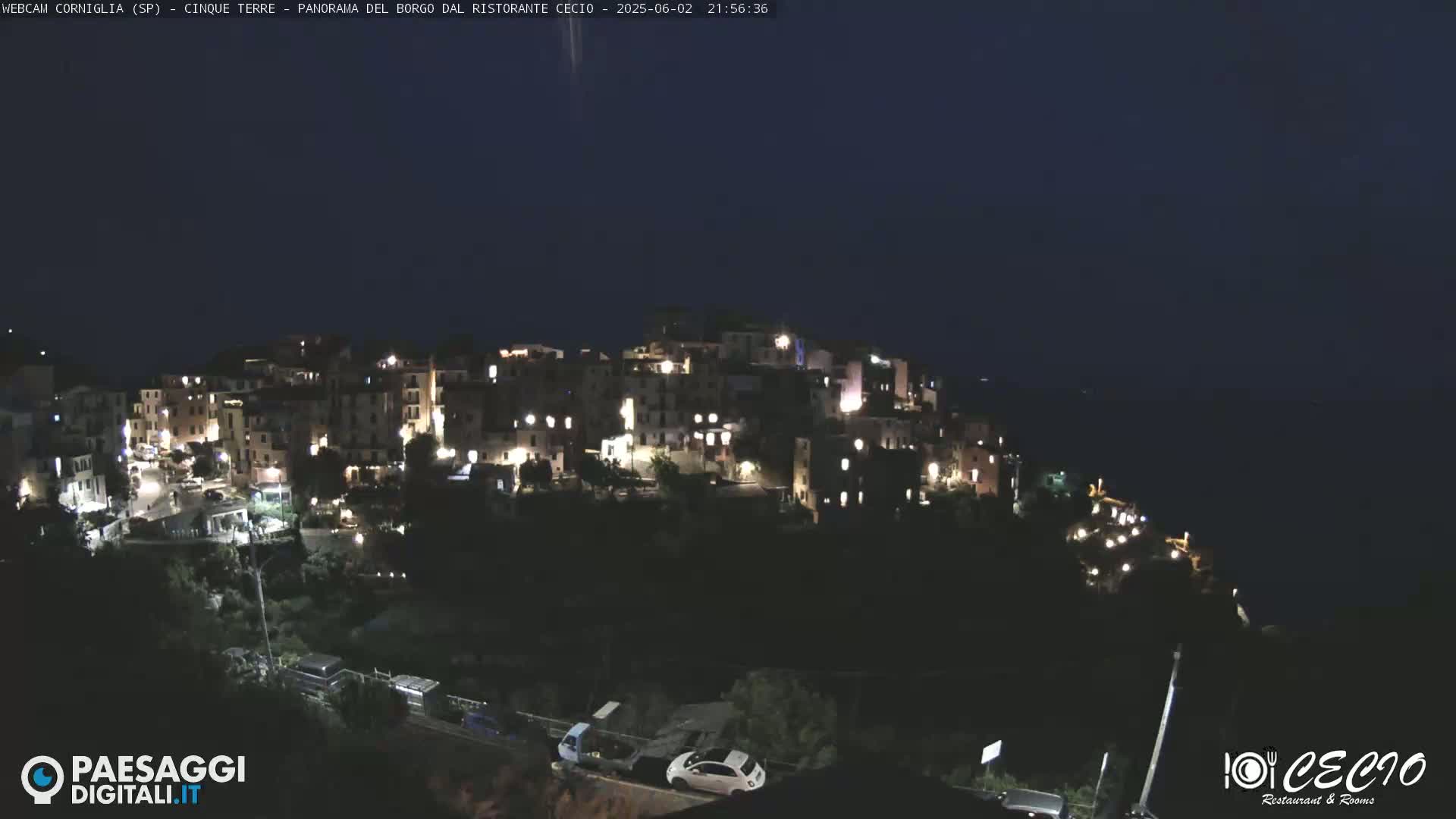 A hillside village at night, illuminated by many windows, overlooks a dark ocean under a clear, dark sky.