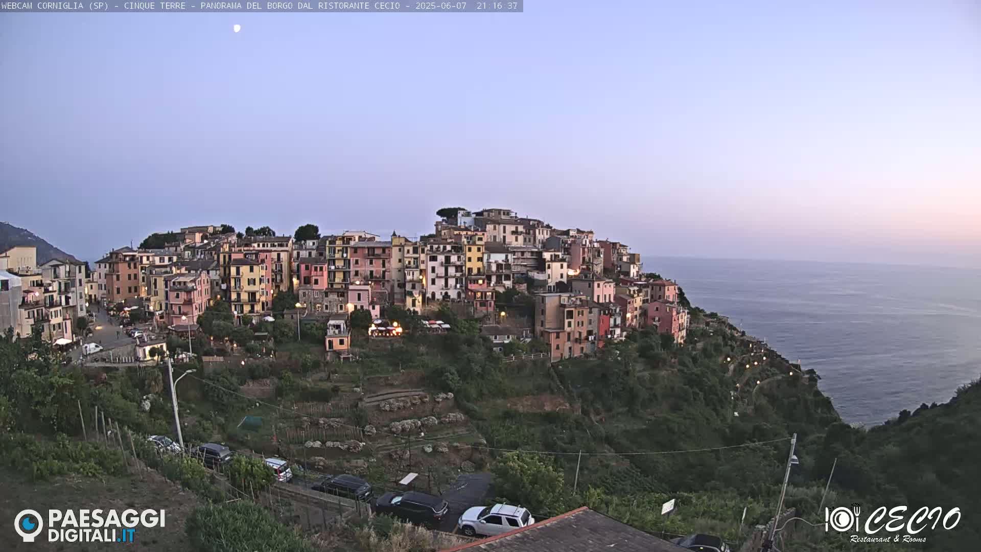 A colorful village clings to a hillside overlooking a calm ocean under a twilight sky.