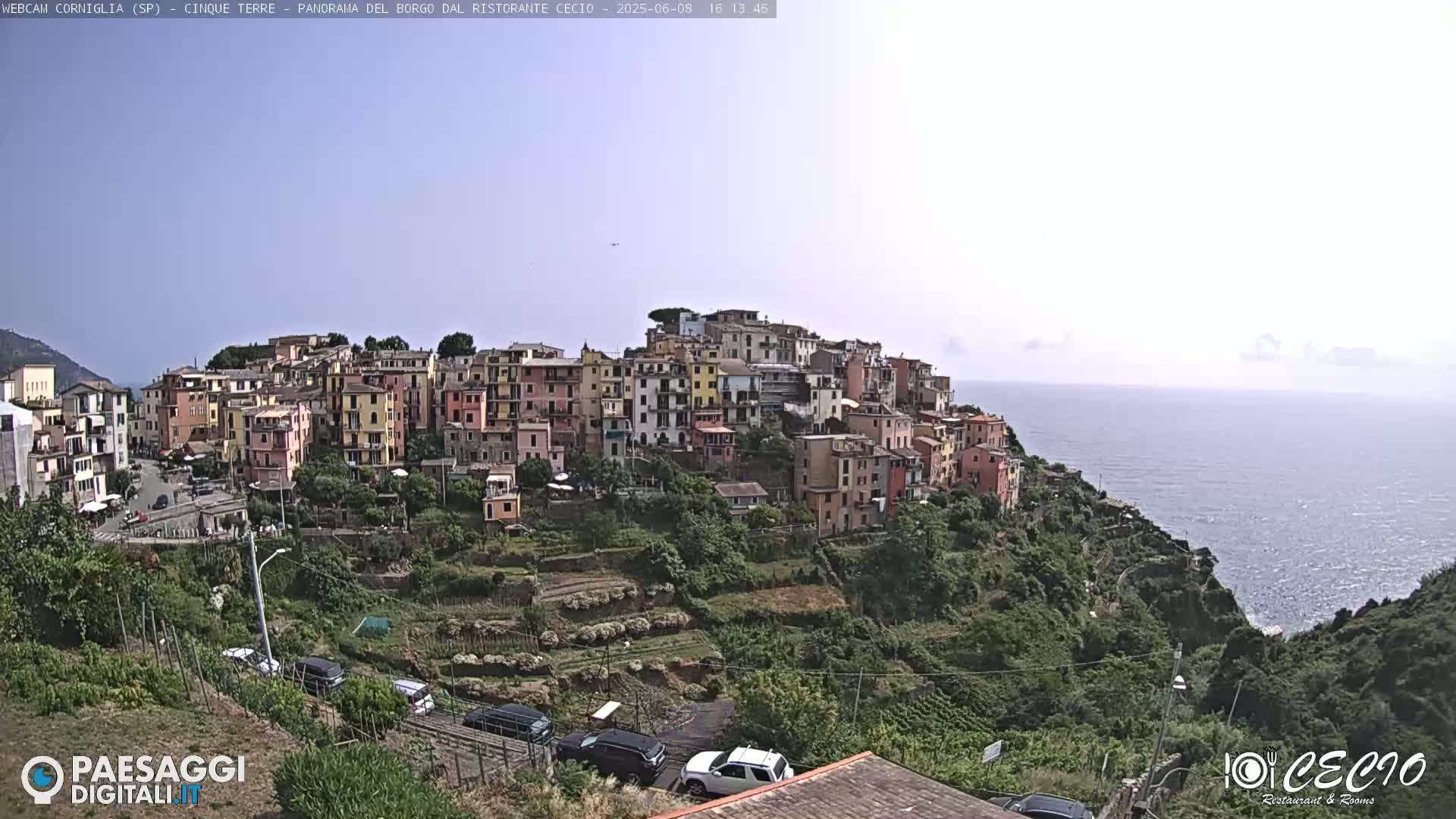 A hillside village of colorful buildings overlooks a calm ocean under a hazy sky.