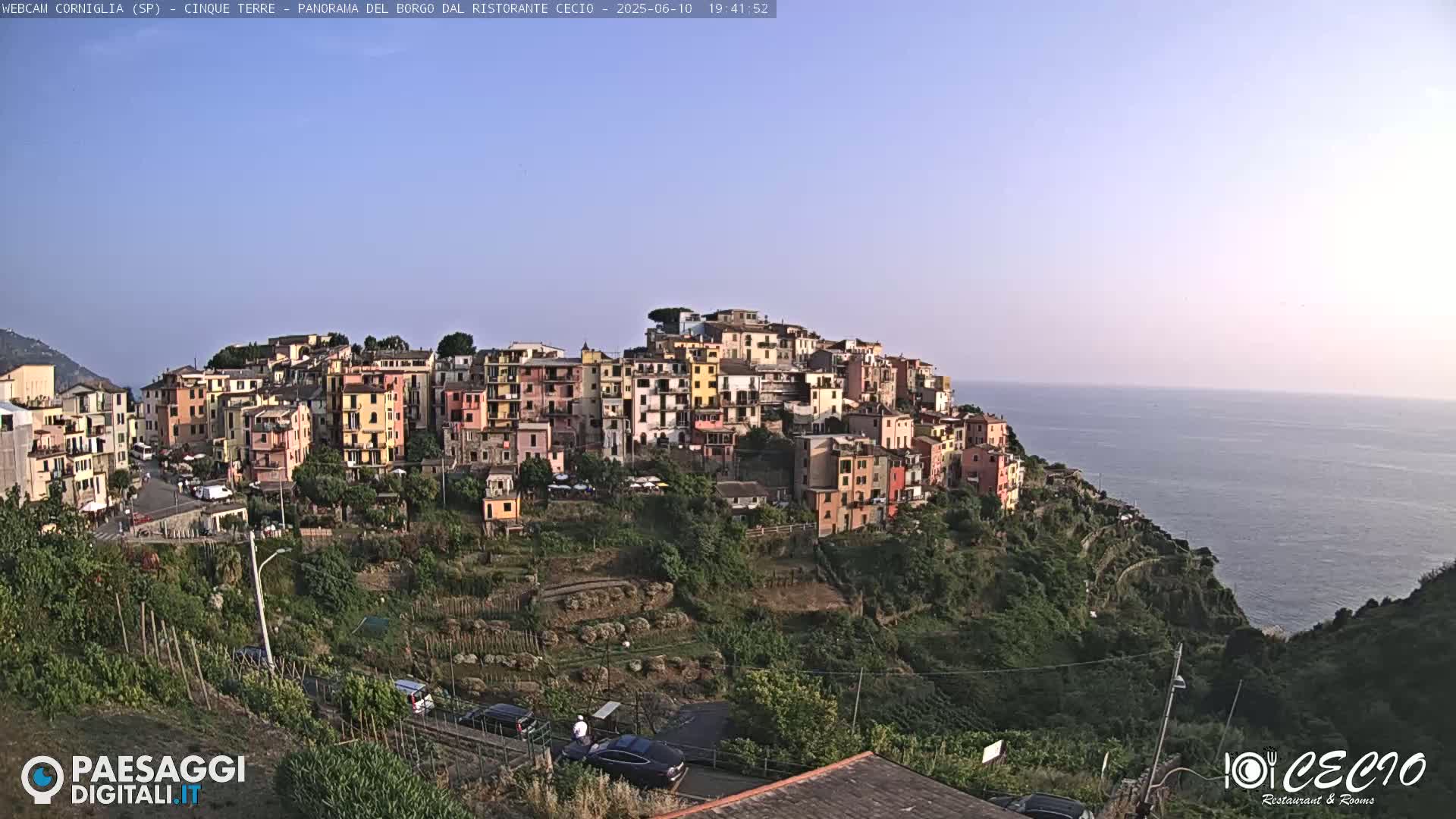 A hillside village of colorful buildings overlooks a calm ocean under a clear, light blue sky.