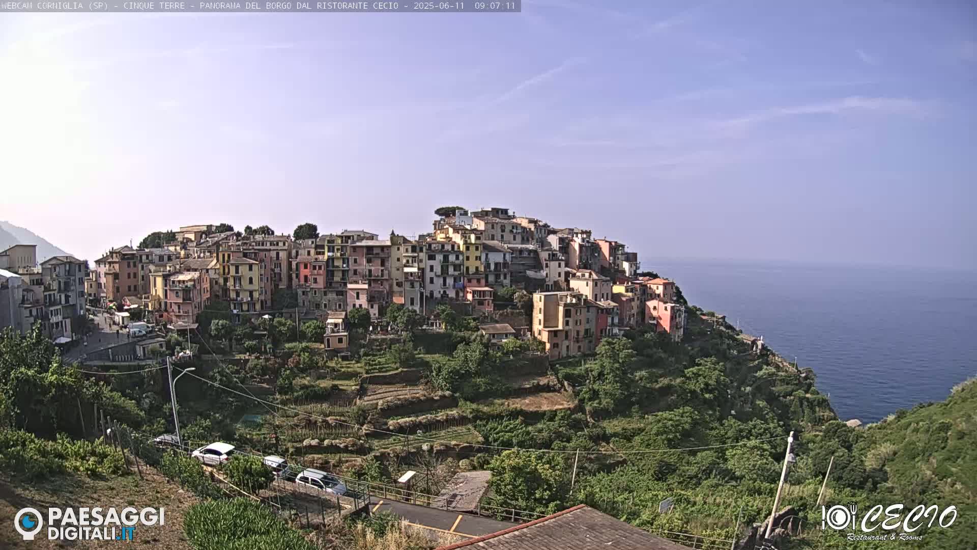 A colorful village clings to a hillside overlooking a calm blue sea under a clear sky.