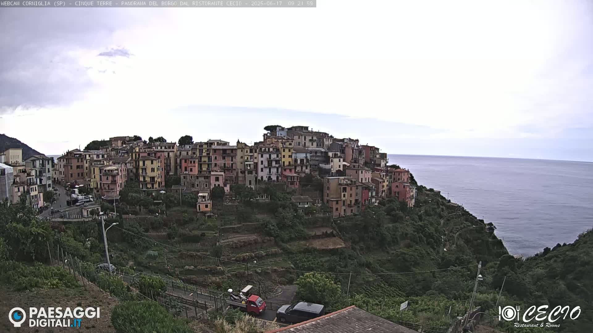 A colorful village clings to a hillside overlooking a calm sea under an overcast sky.
