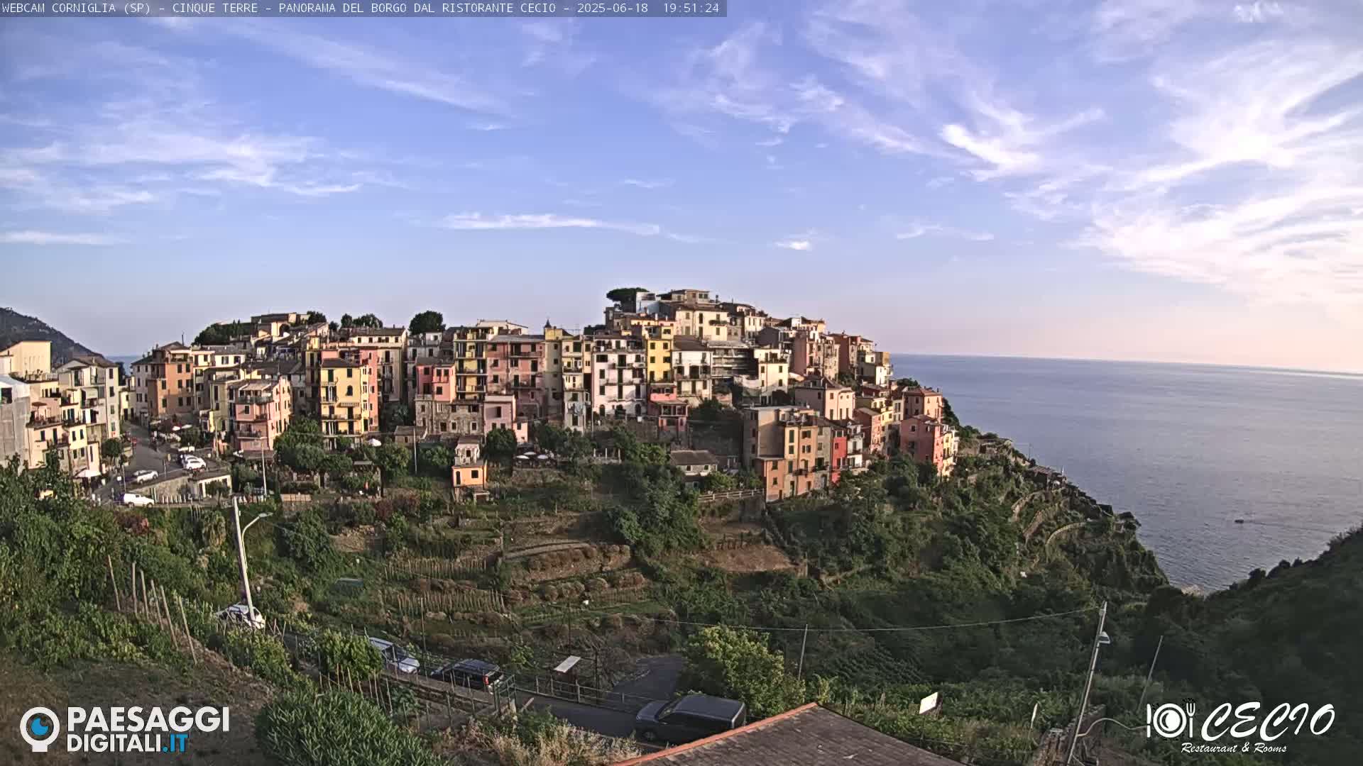 A picturesque village of colorful buildings clings to a hillside overlooking a calm sea under a mostly clear sky.
