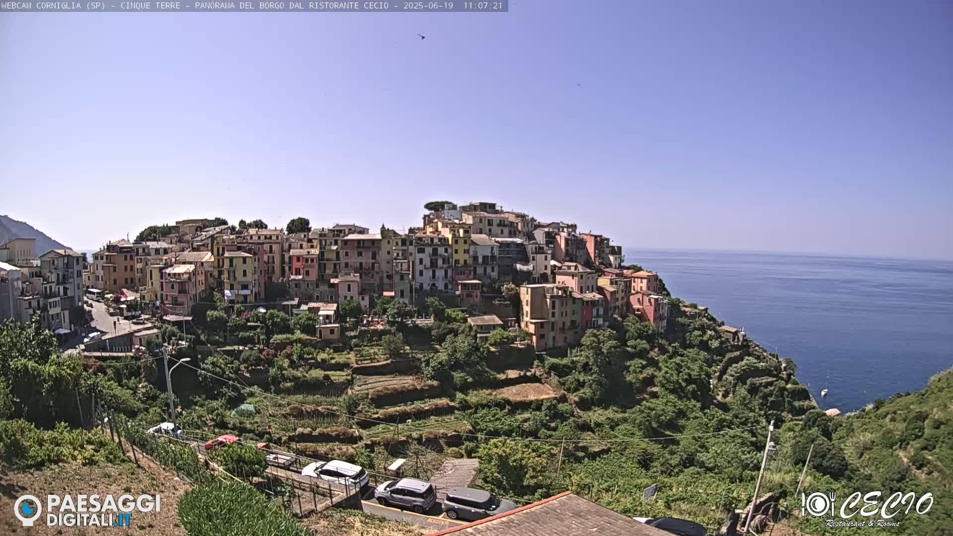 A colorful village clings to a hillside overlooking a calm, blue sea under a clear sky.
