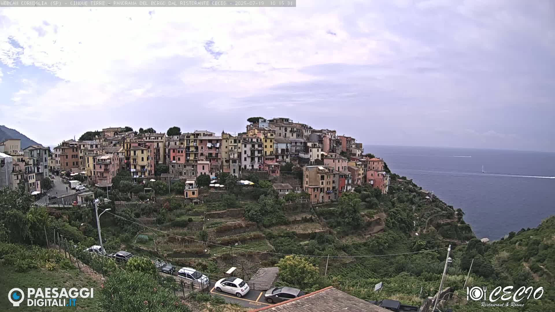 A colorful hillside village overlooks a calm ocean under a mostly cloudy sky.