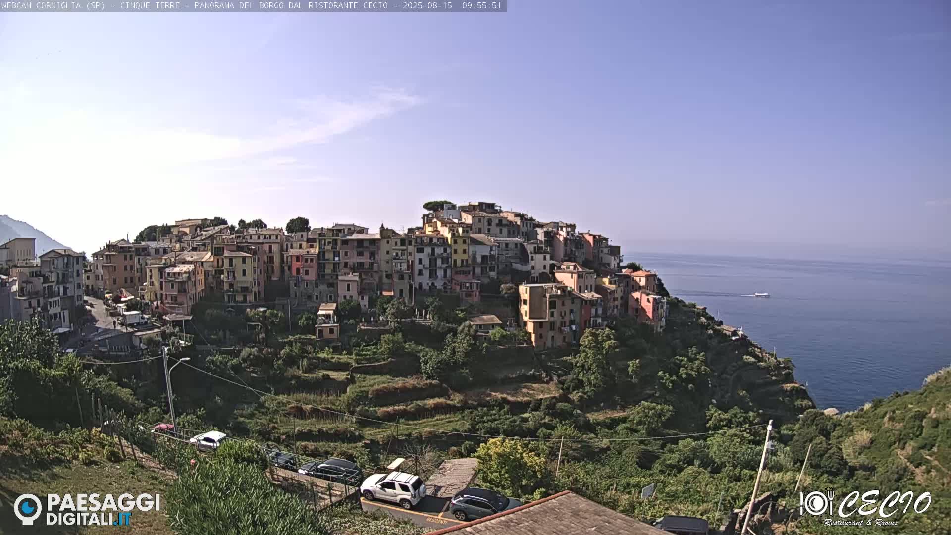 A picturesque village of colorful buildings clings to a hillside overlooking a calm sea under a clear blue sky.