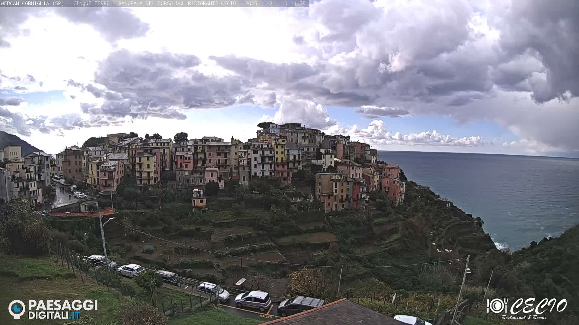 A vibrant coastal village with colorful buildings is nestled on a terraced green hillside overlooking a deep blue, slightly choppy sea, under a dynamic sky featuring a mix of bright white and dark grey clouds.