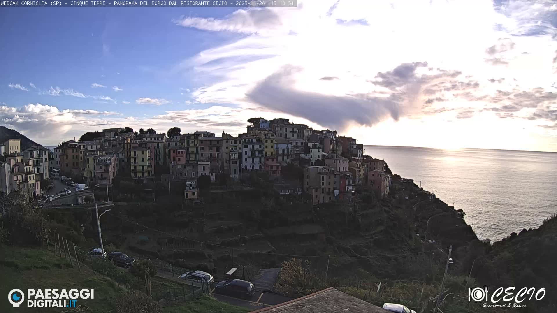 A vibrant coastal village with colorful buildings is nestled on a terraced green hillside overlooking a deep blue, slightly choppy sea, under a dynamic sky featuring a mix of bright white and dark grey clouds.