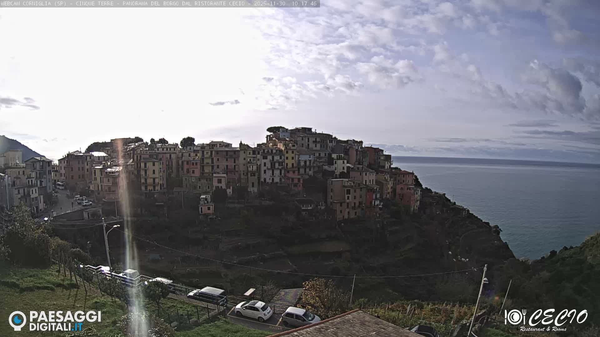 A vibrant coastal village with colorful buildings is nestled into a steep, terraced hillside above a calm sea under a partially sunny sky with scattered clouds.