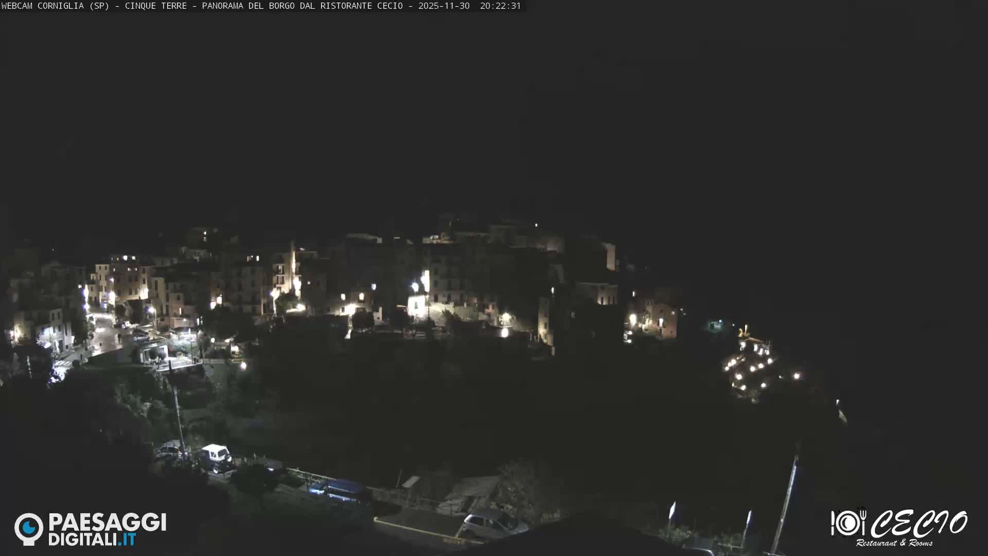 A vibrant coastal village with colorful buildings is nestled into a steep, terraced hillside above a calm sea under a partially sunny sky with scattered clouds.