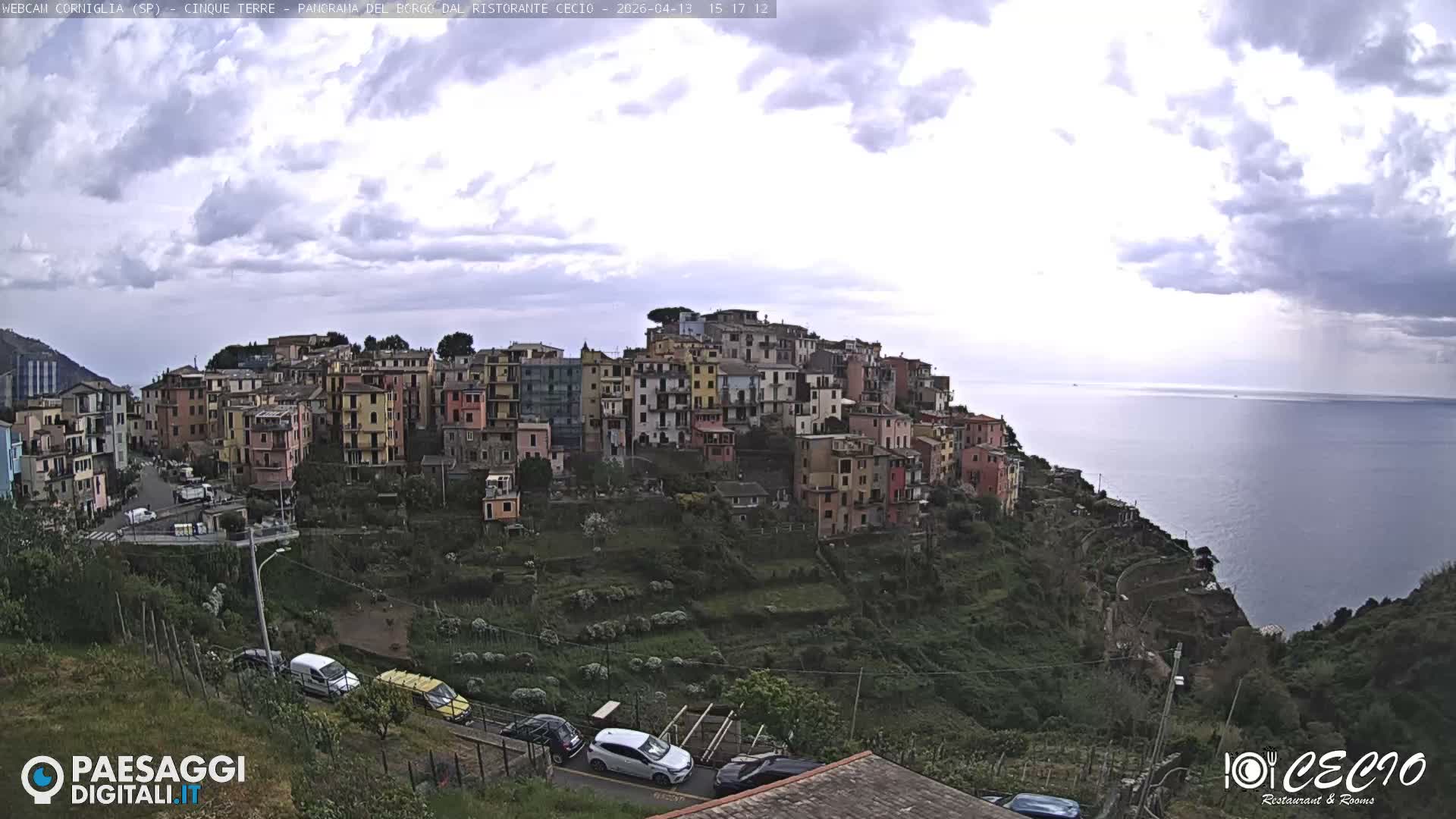 A vibrant, colorful village is densely built into a steep, terraced hillside overlooking a calm sea under a cloudy, overcast sky.