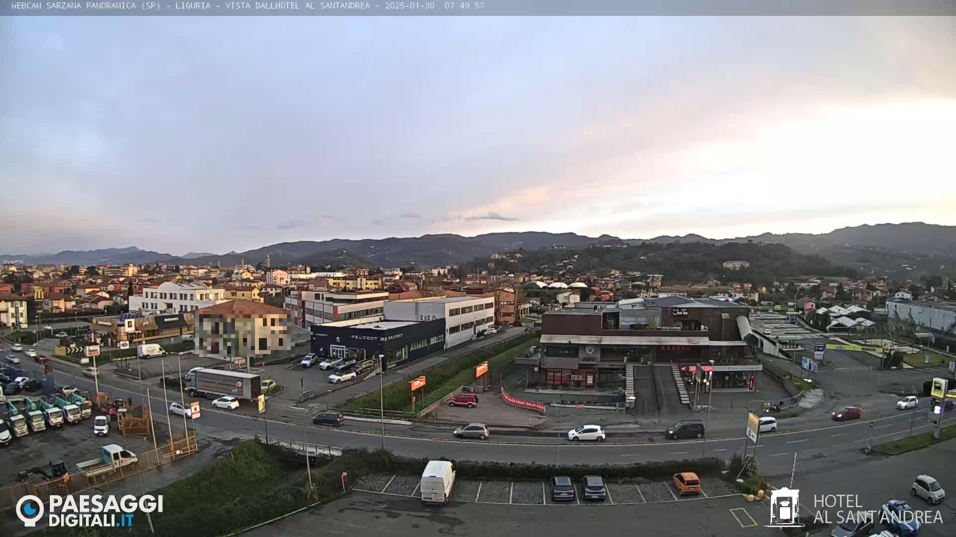 A wide shot of a town under a pale, overcast sky shows buildings, roads, and parked cars, with hills in the background.