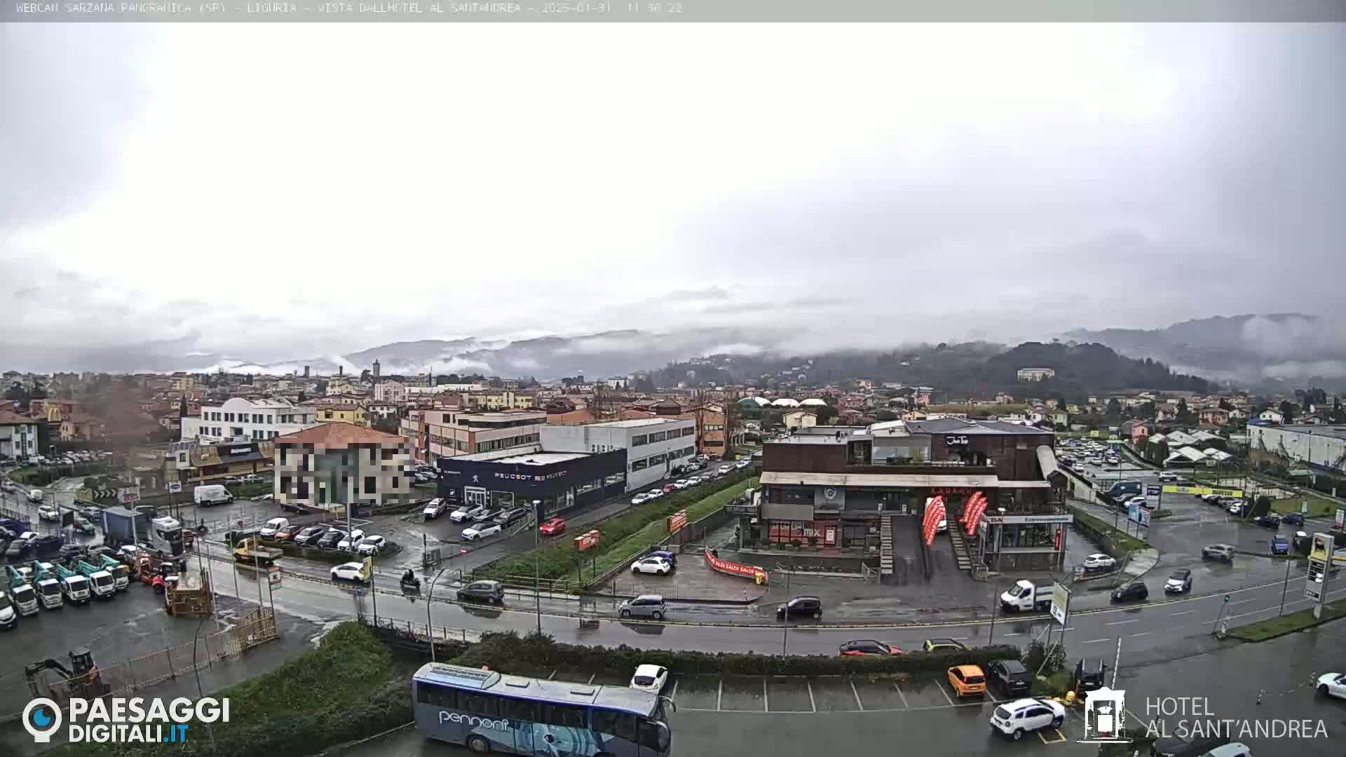 A misty, overcast day reveals a town's cityscape, featuring buildings, parking lots filled with cars, and a bus, all situated near a mountain range shrouded in fog.