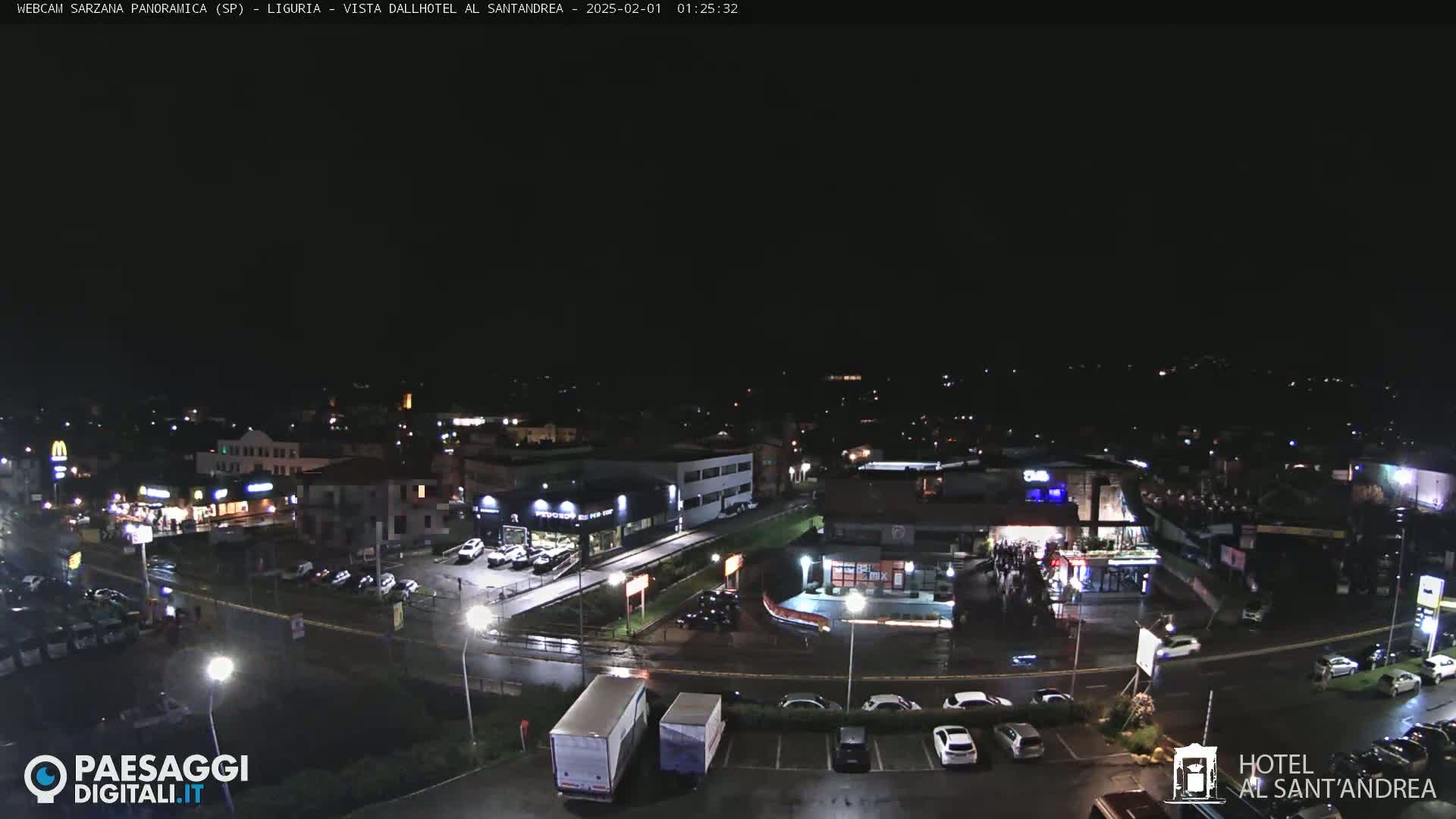 A nighttime, wet aerial view of a town with illuminated buildings, roads, and parked cars.