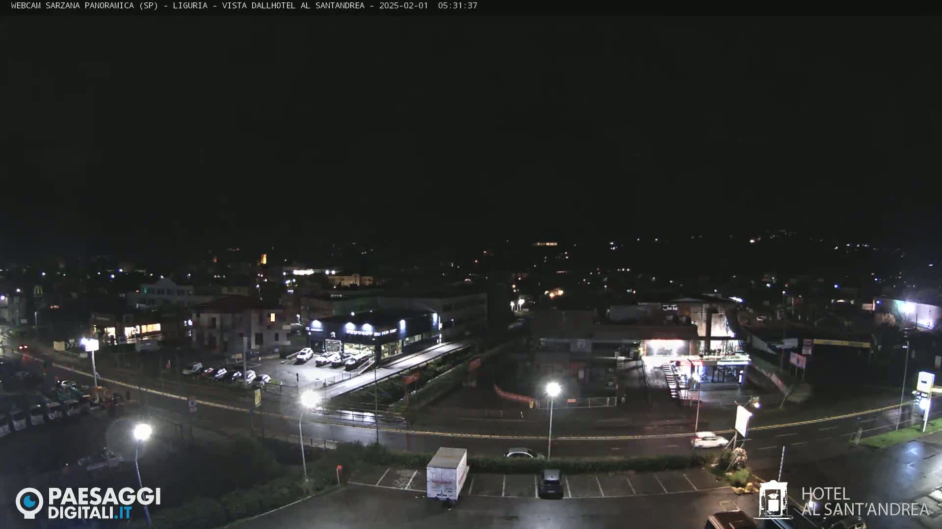 A nighttime, wet, aerial view of a town with streetlights illuminating cars in parking lots and buildings along roads.