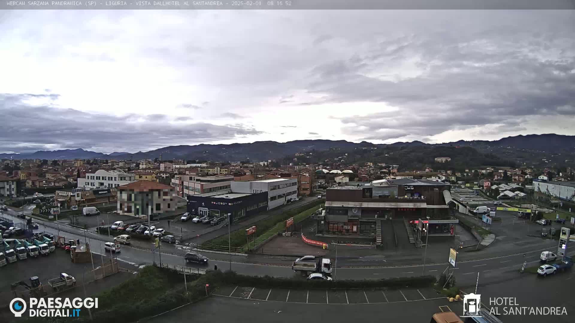 A panoramic view of a town under a cloudy sky shows buildings, roads, cars, and distant mountains.
