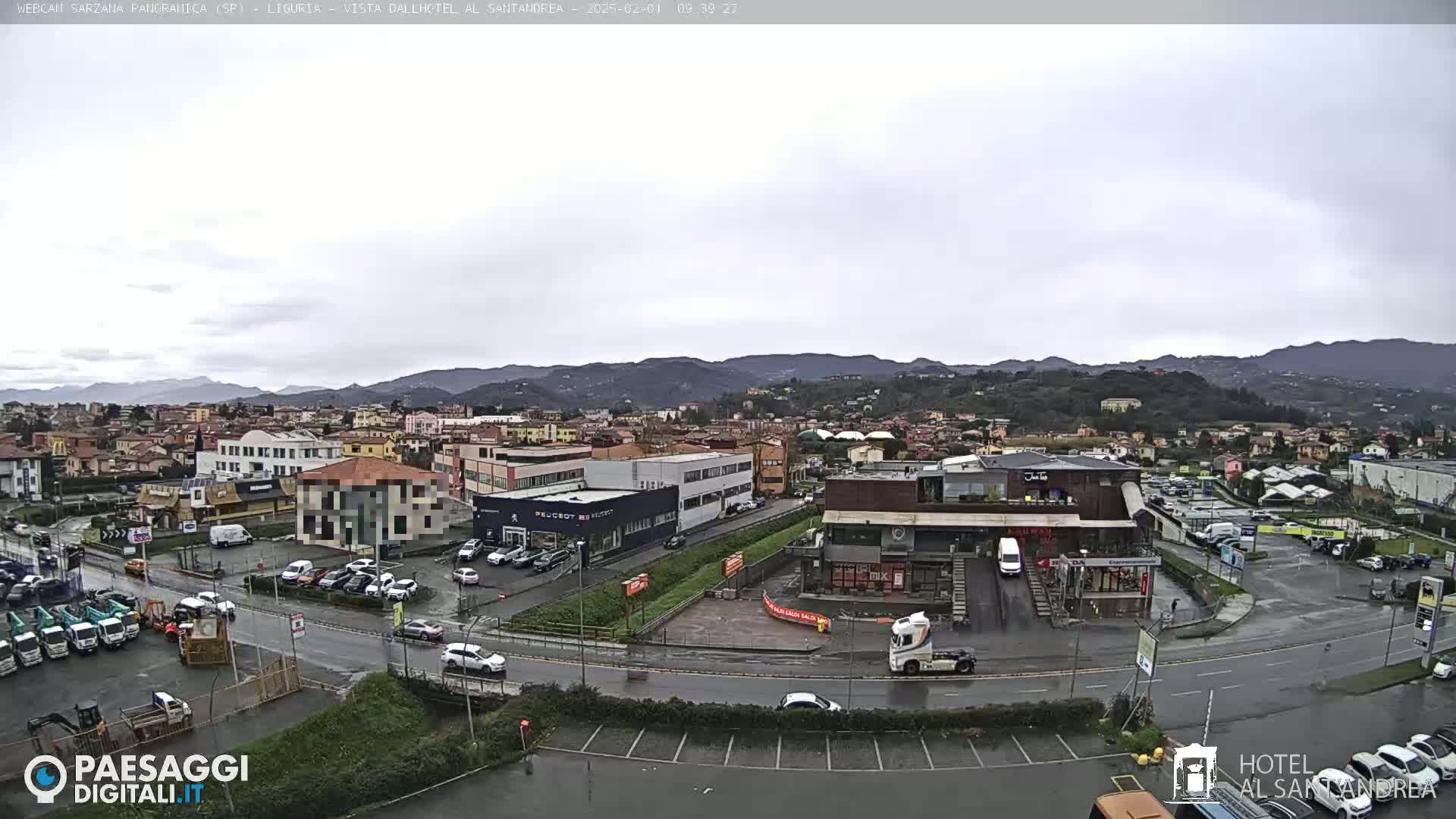 A wet, overcast day reveals a sprawling town nestled against a backdrop of rolling hills, with various buildings, parked cars, and construction equipment visible in a large parking lot.