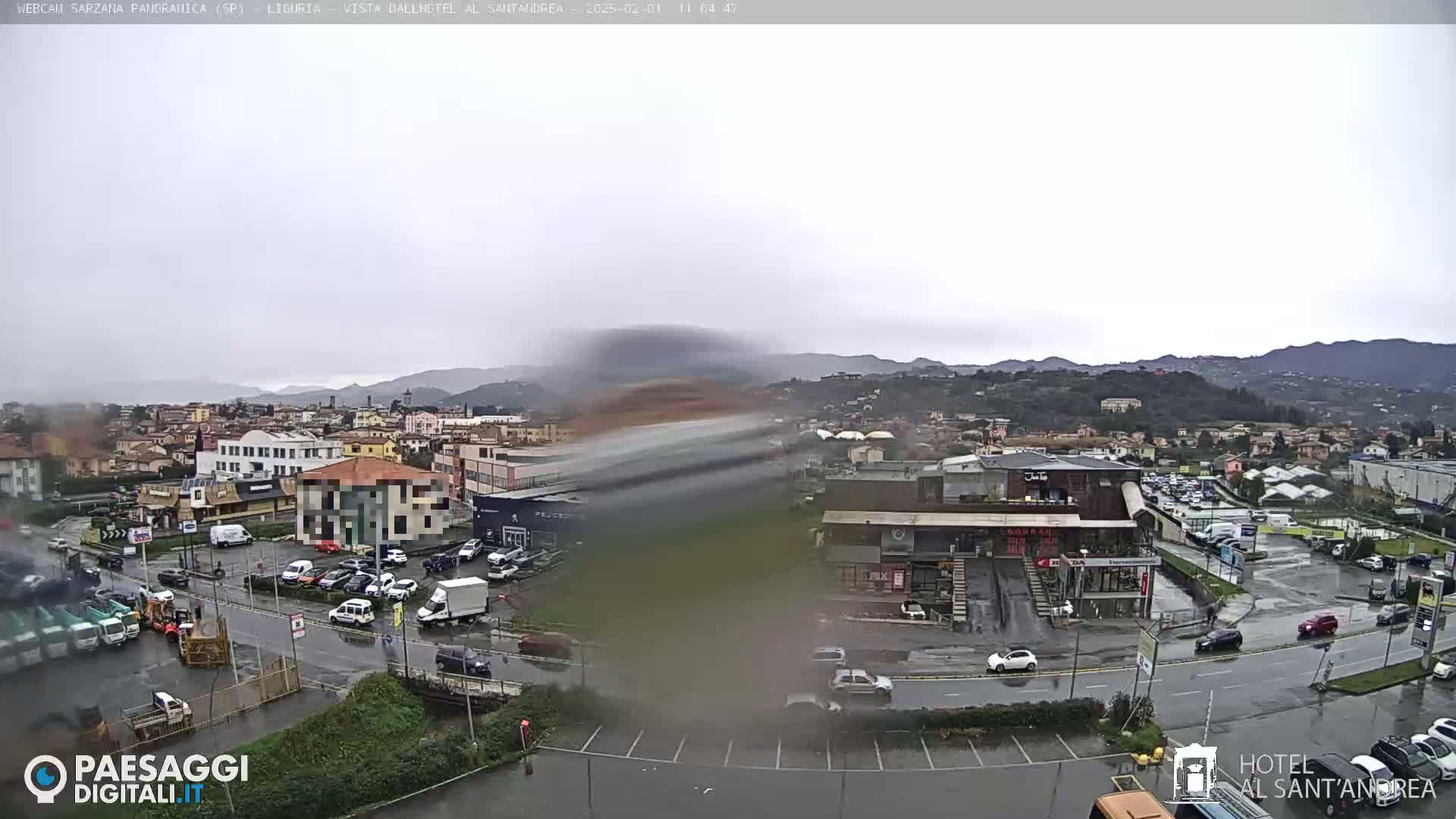 A rain-soaked town with cars parked in lots and driving on wet streets, nestled among hills under a cloudy sky.