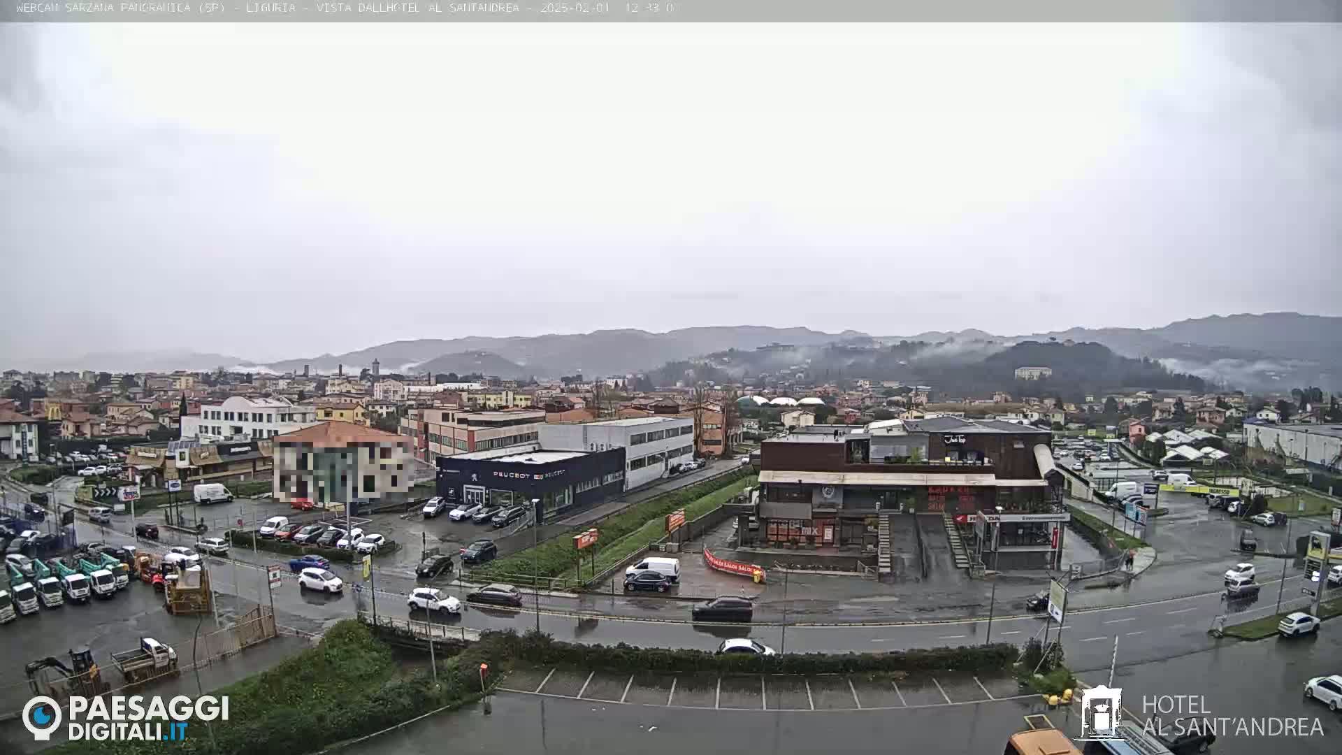 A wet, overcast view of a town's outskirts, showing buildings, parking lots with cars, and distant hills shrouded in mist.