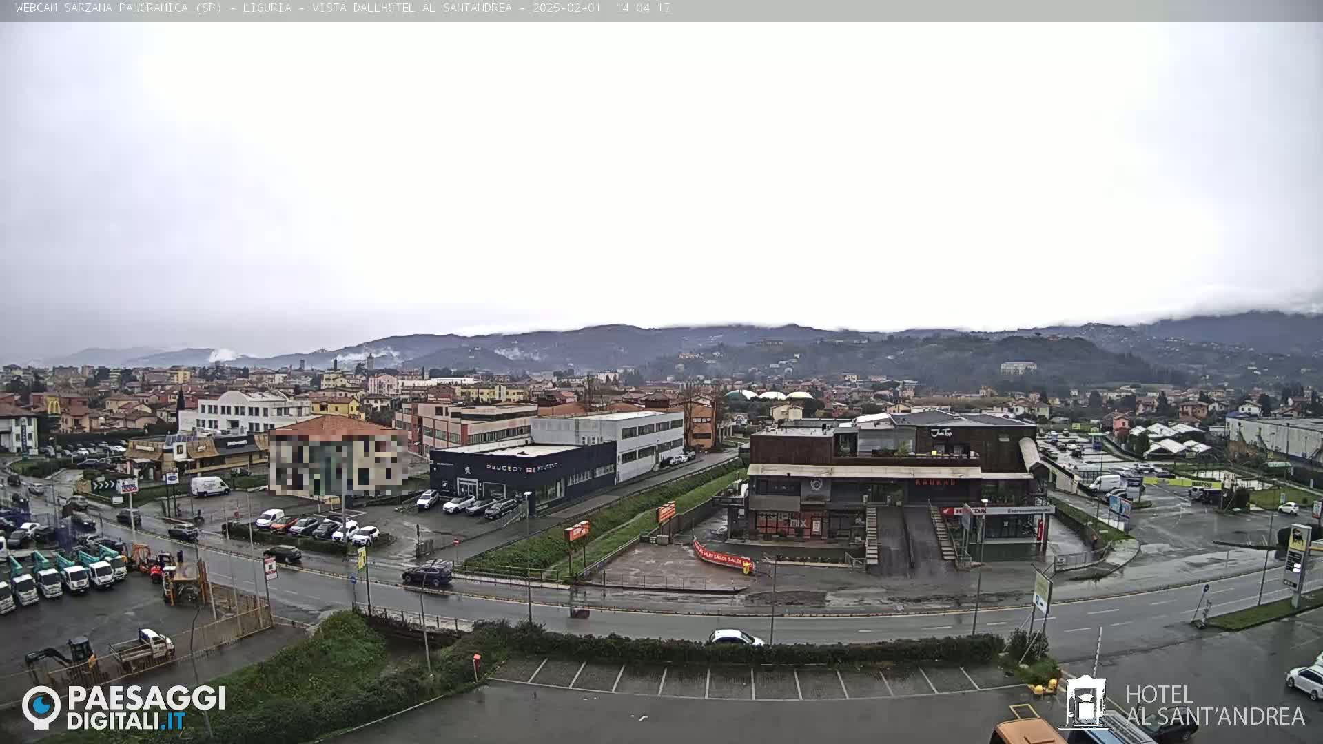 A wide shot of a town under an overcast sky shows a variety of buildings, including industrial structures and businesses, with wet roads and parking lots, surrounded by hills.