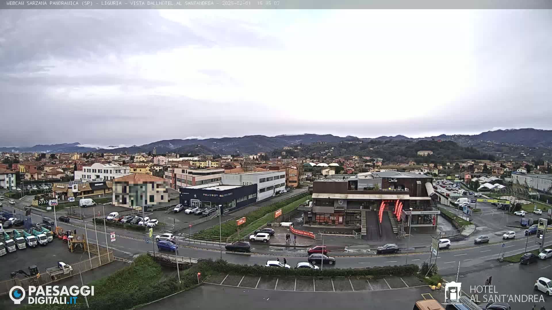 A view of a town and surrounding hills under an overcast sky, showing roads, buildings, cars, and a parking lot.
