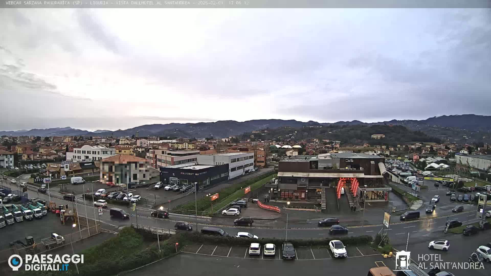 A cloudy day overlooks a town with buildings, roads, and parked cars, mountains in the distance.