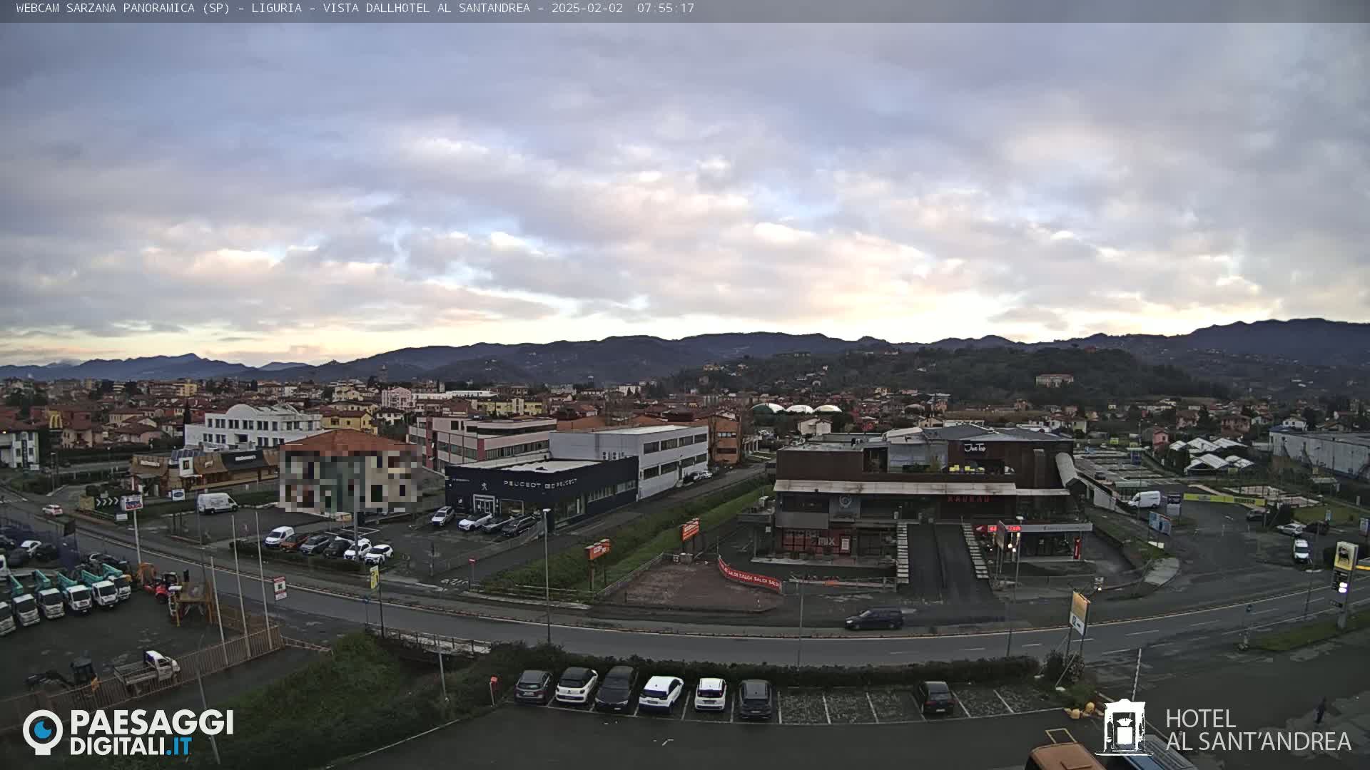 A wide shot of a town at sunrise under a mostly cloudy sky, showing buildings, roads, cars, and distant mountains.