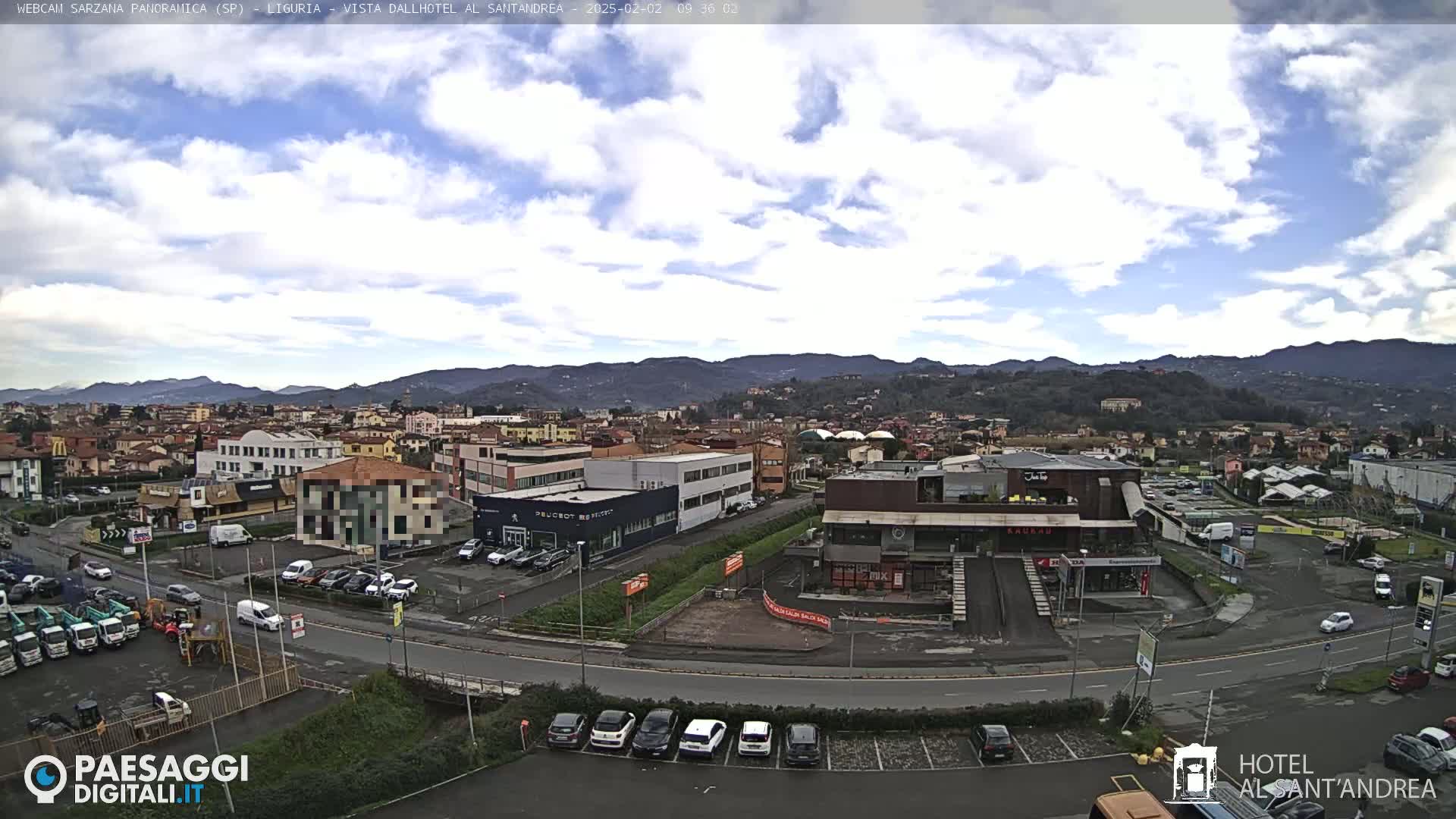 A partly cloudy day reveals a sprawling town nestled amongst hills, with various buildings, parking lots, and vehicles visible from a high vantage point.