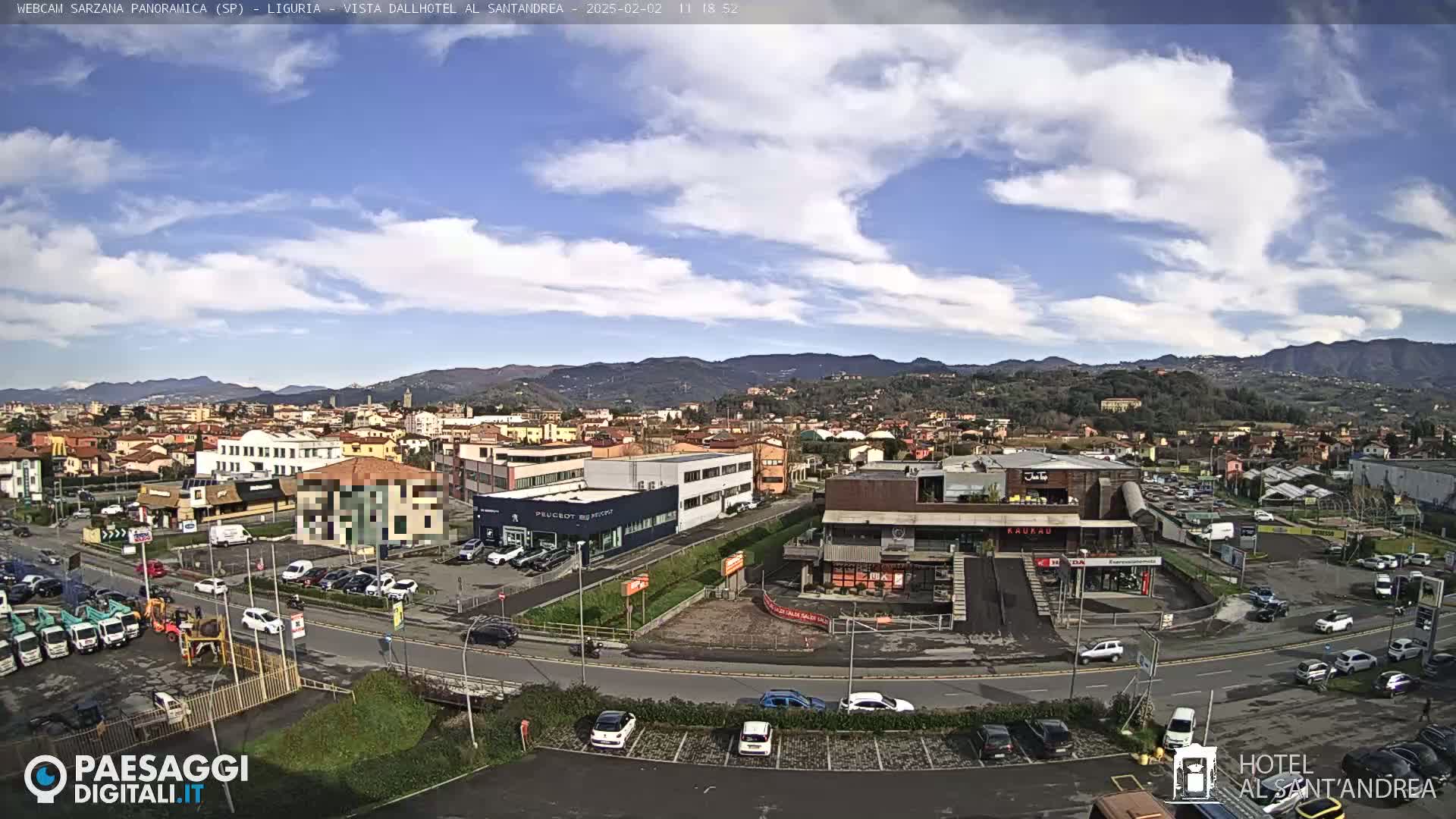 A panoramic view of a town nestled in a valley, under a partly cloudy sky, shows a mix of residential and commercial buildings, with cars parked in lots and driving on roads.