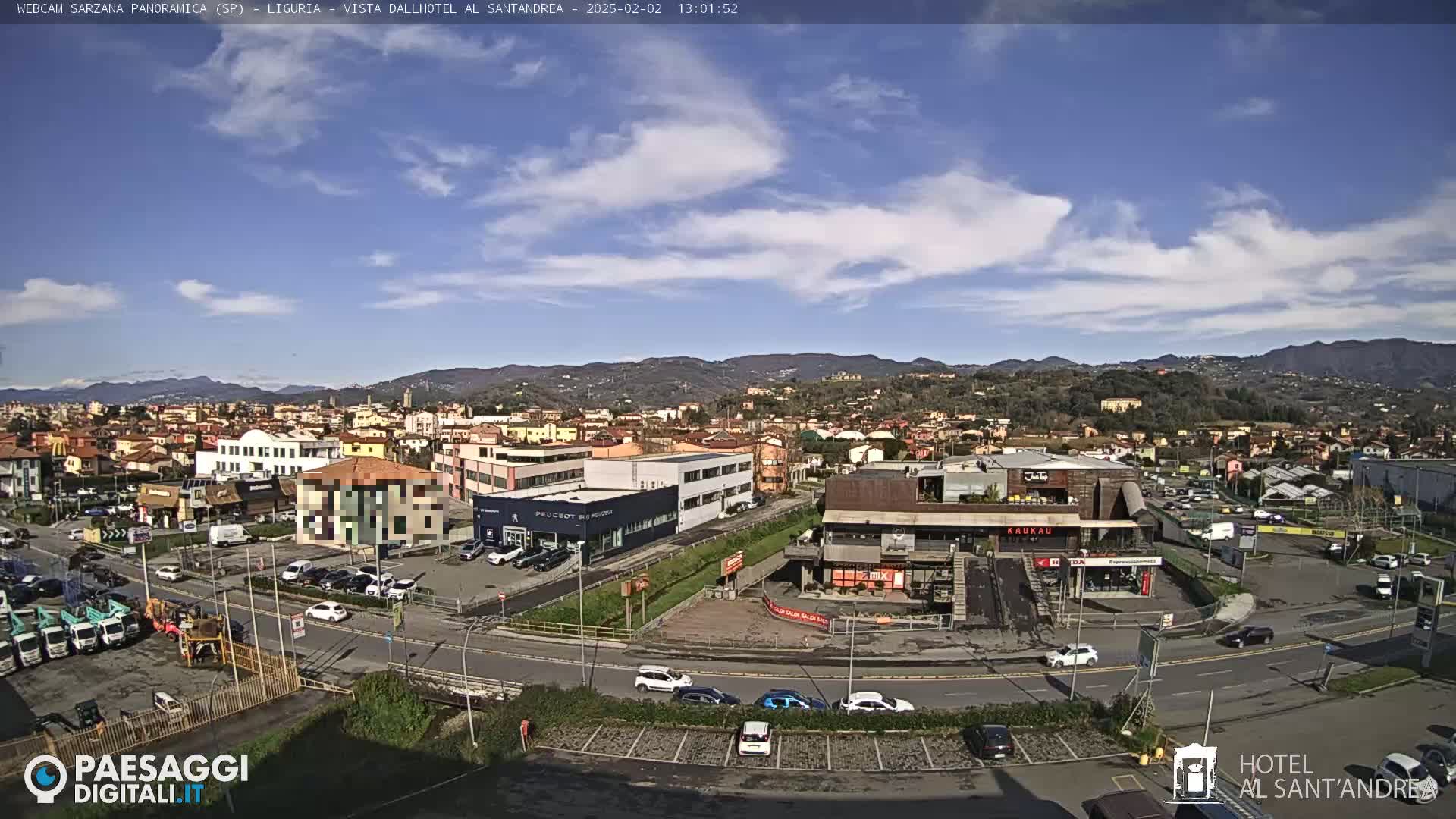 A panoramic view of a town on a partly sunny day, showing buildings, roads, and parked cars, with hills and mountains in the background.