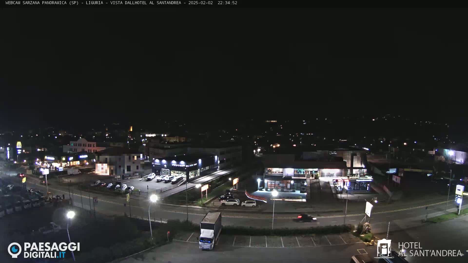 A nighttime, aerial view of a town's outskirts, showing roads, buildings, and parked cars under a clear, dark sky.