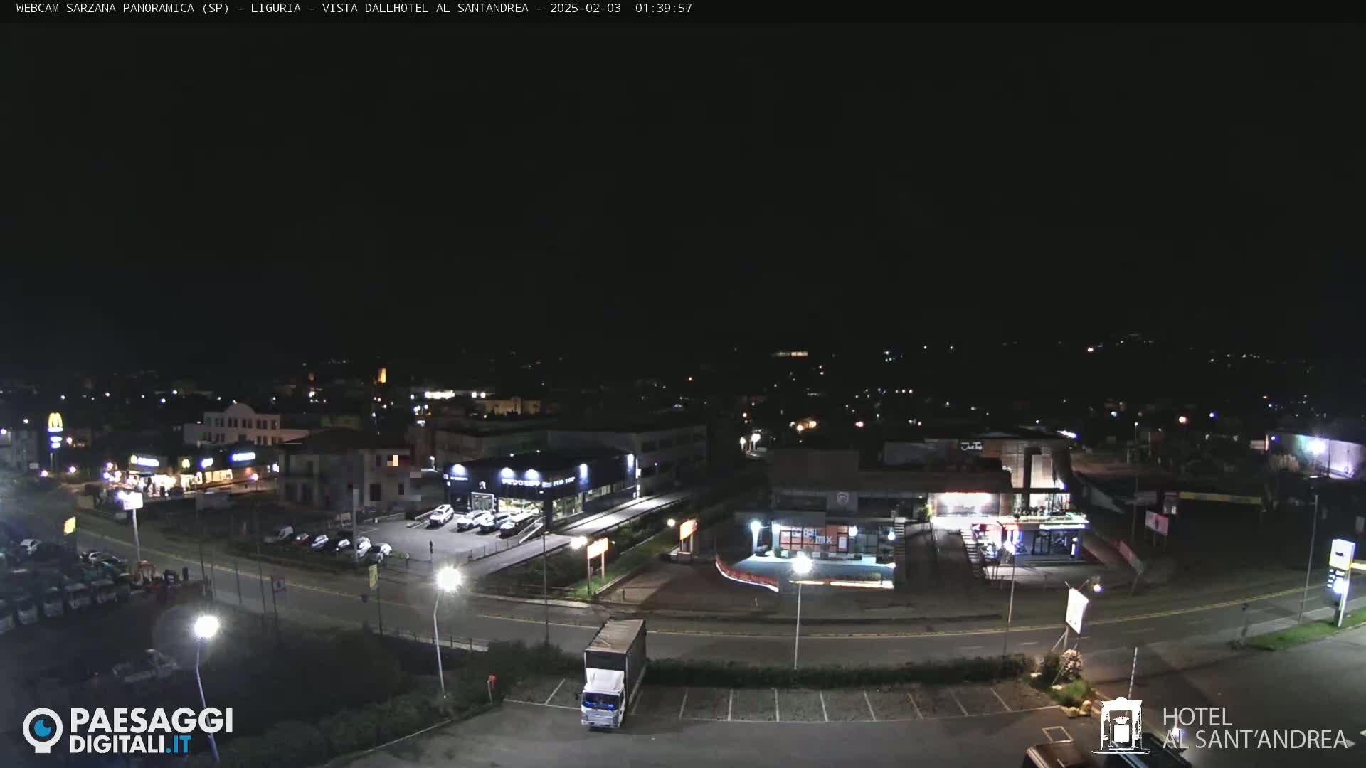 A nighttime, aerial view of a town’s outskirts showing a road, parking lots, and various businesses illuminated by artificial lights.