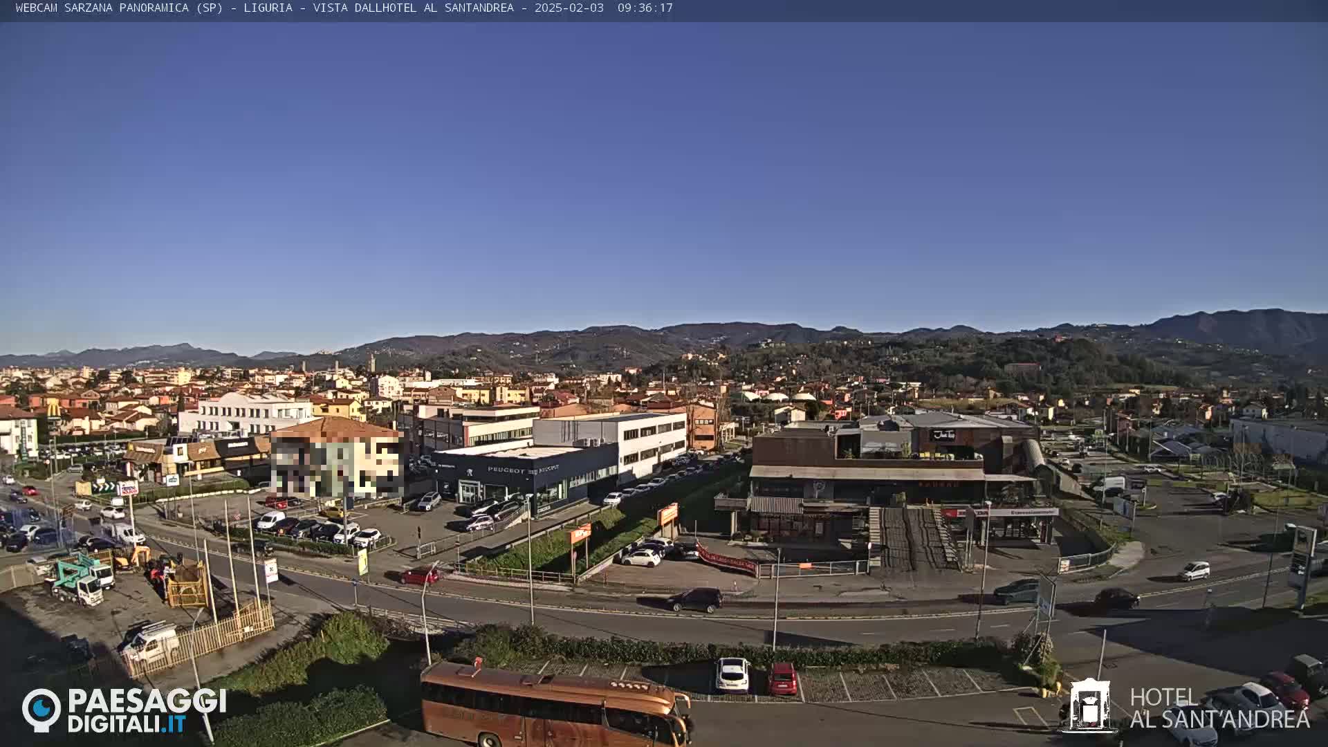 A clear, sunny day reveals a sprawling town nestled amongst rolling hills, with a variety of buildings, vehicles, and a large bus visible in a parking lot.