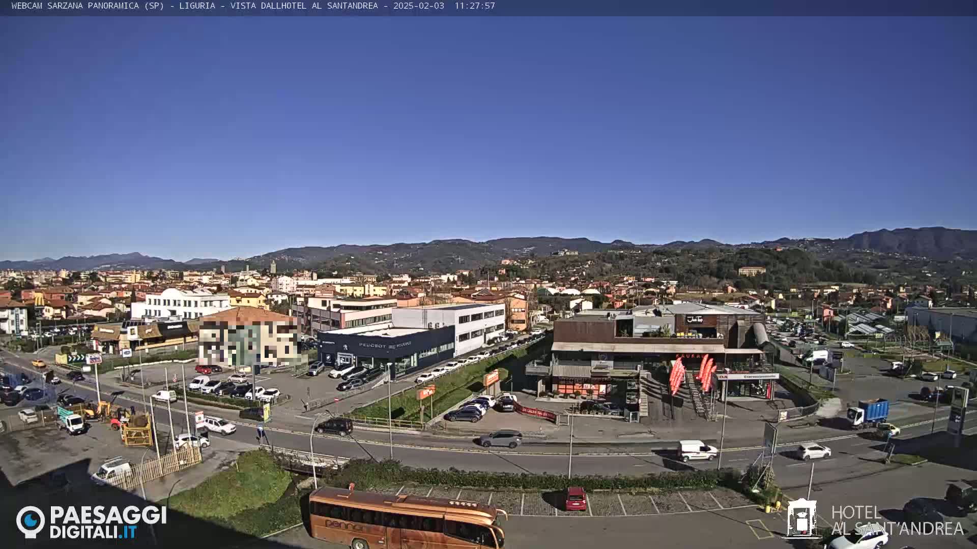 A wide, sunny outdoor view of a town with buildings, cars, and a large bus in a parking lot, mountains visible in the background.