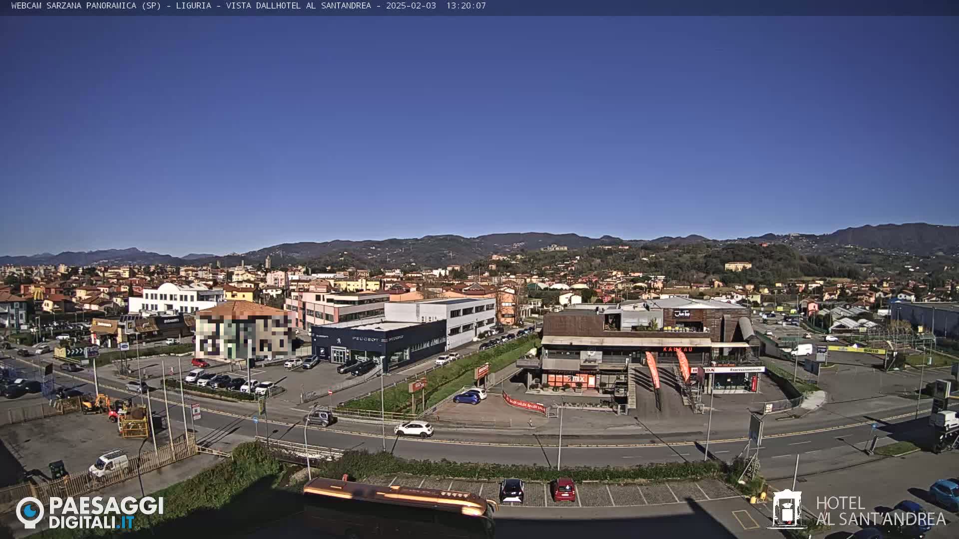 A panoramic view of a town nestled in a valley under a clear blue sky, with buildings, roads, and parked cars visible.