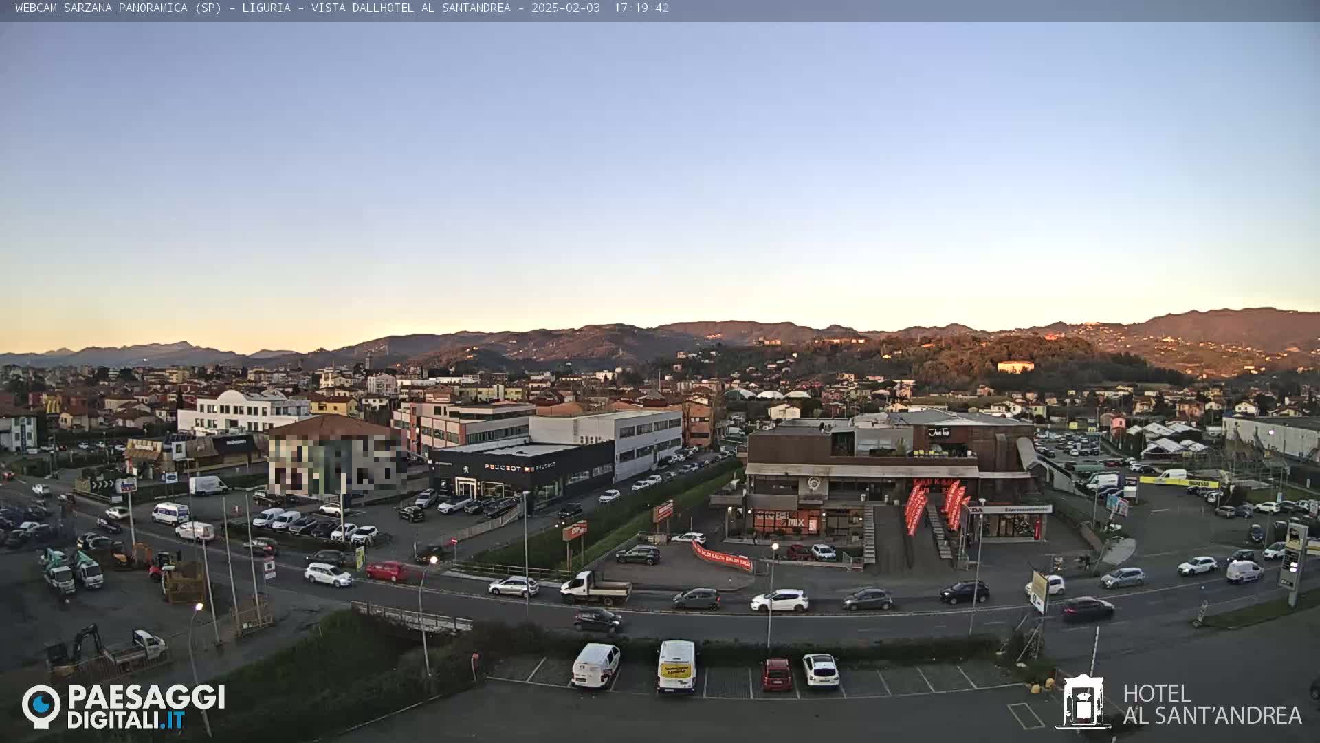 A clear evening view of a town nestled in a valley, surrounded by hills, with cars parked in lots and driving on roads.