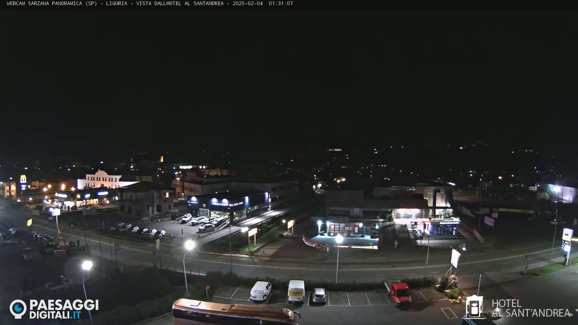 A nighttime, high-angle view of a town's street and parking lots with various buildings, including restaurants and a car dealership, illuminated by streetlights.