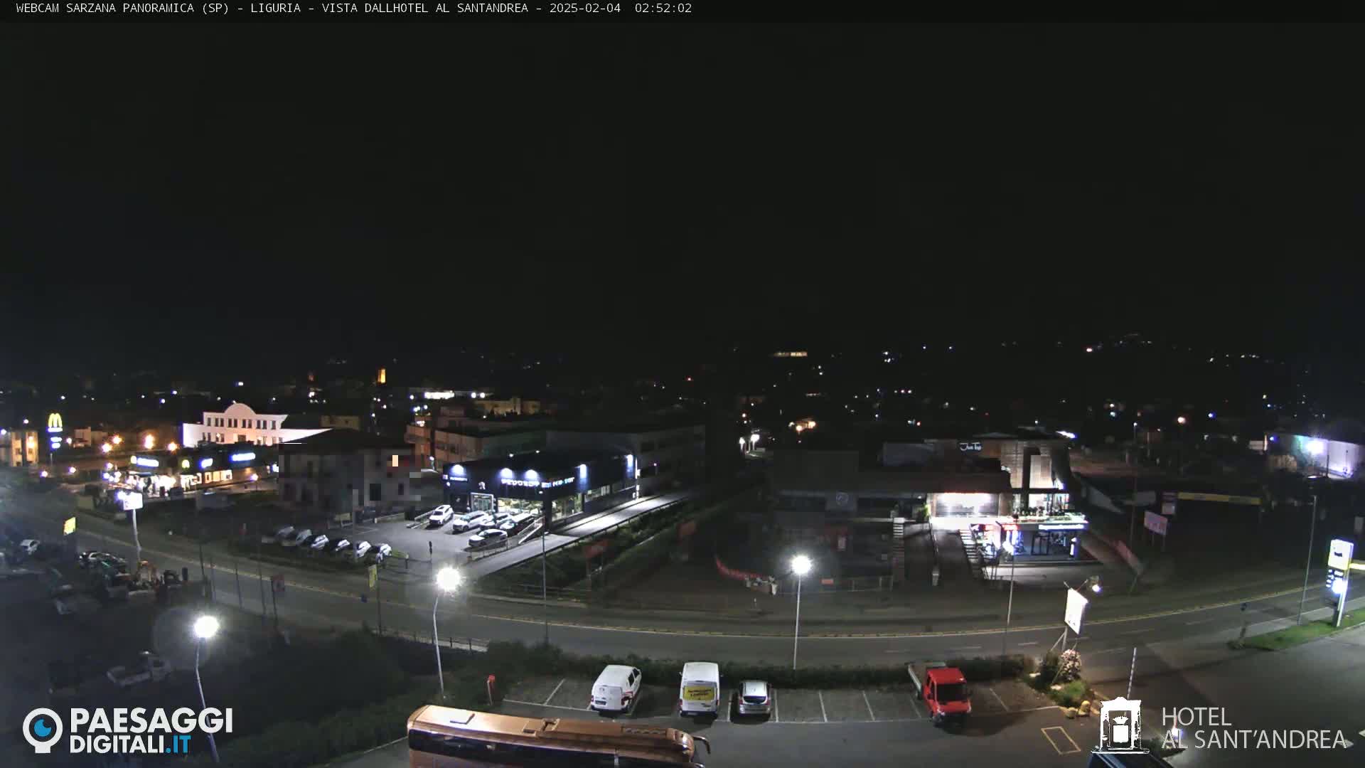 A nighttime, aerial view of a town's outskirts, showing a road curving around businesses and parking lots with cars.