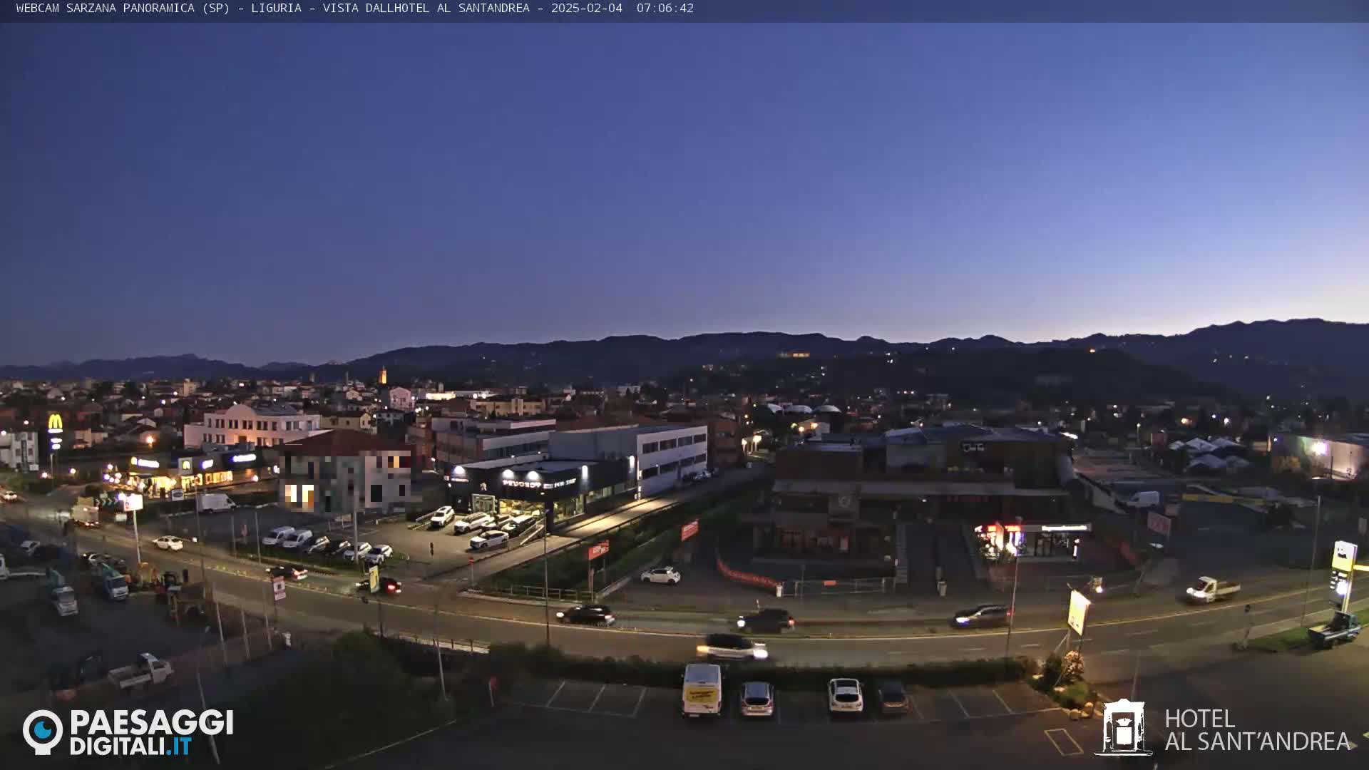 A twilight view of a town nestled beside mountains, with cars moving along roads and a few lights visible in the buildings.