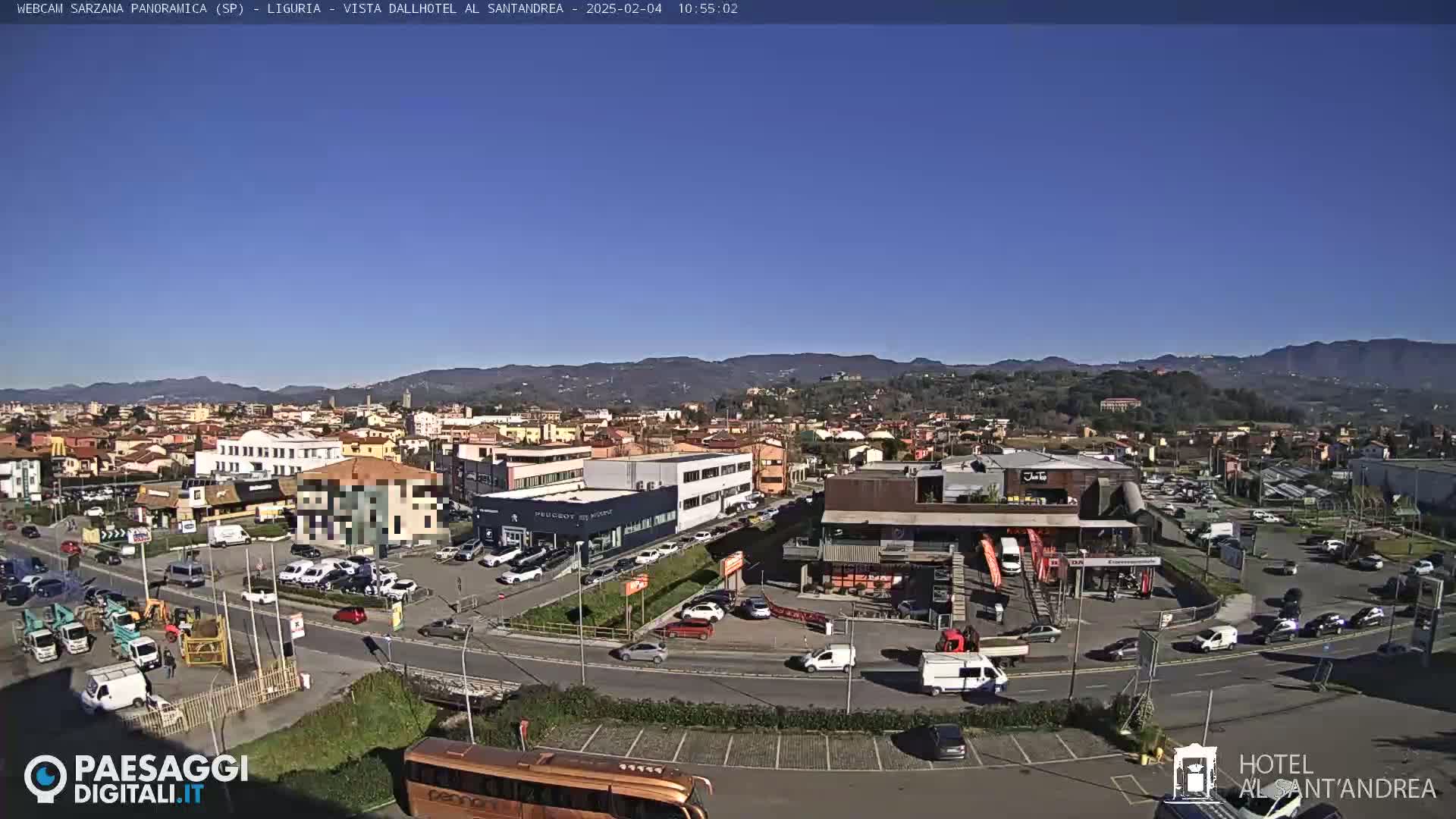 A clear, sunny day reveals a sprawling cityscape nestled against a backdrop of rolling hills, with numerous cars parked in lots and traffic moving along the roads.